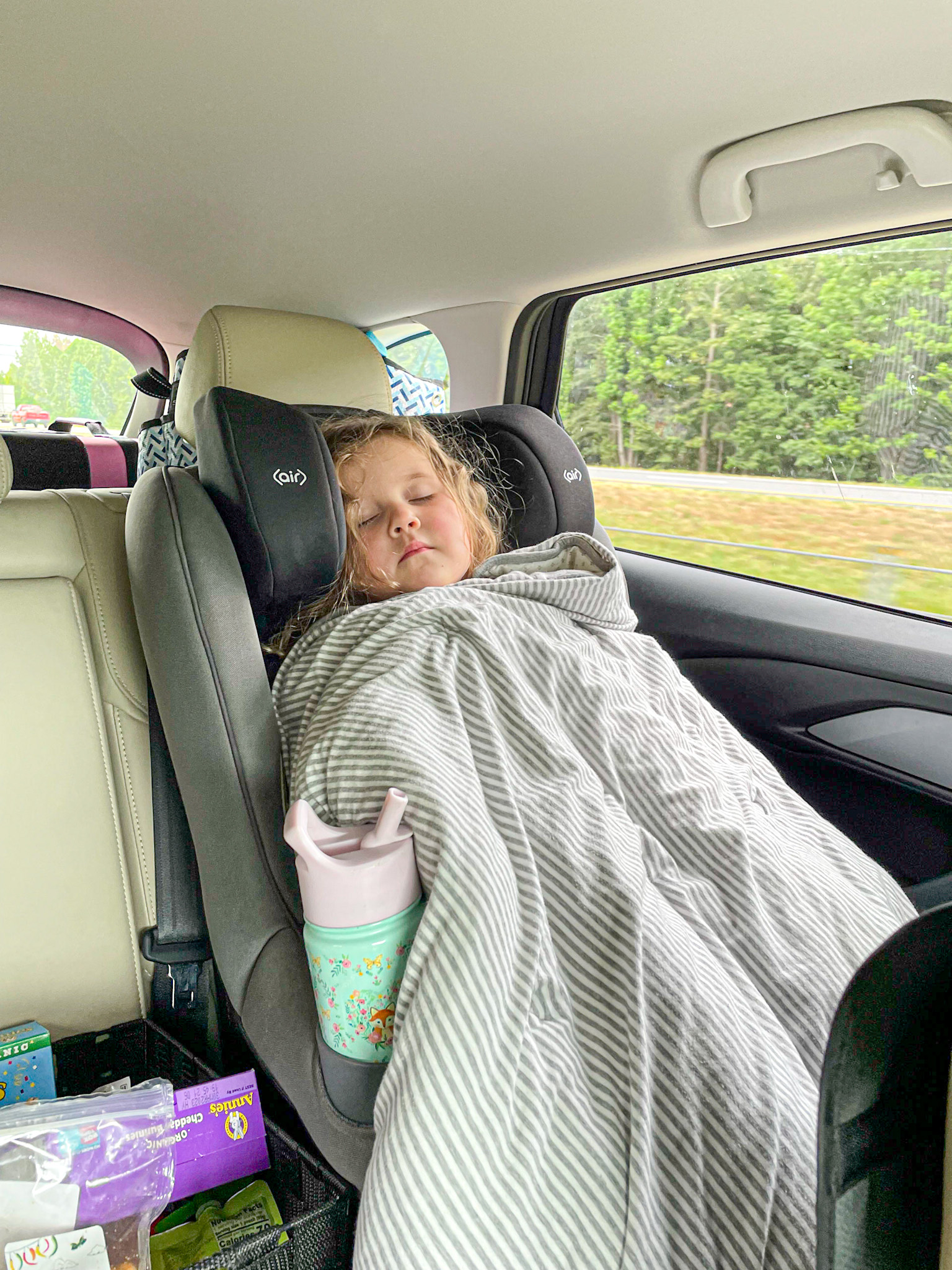 A toddler girl is covered with a blanket sleeping in her car seat during a road trip.