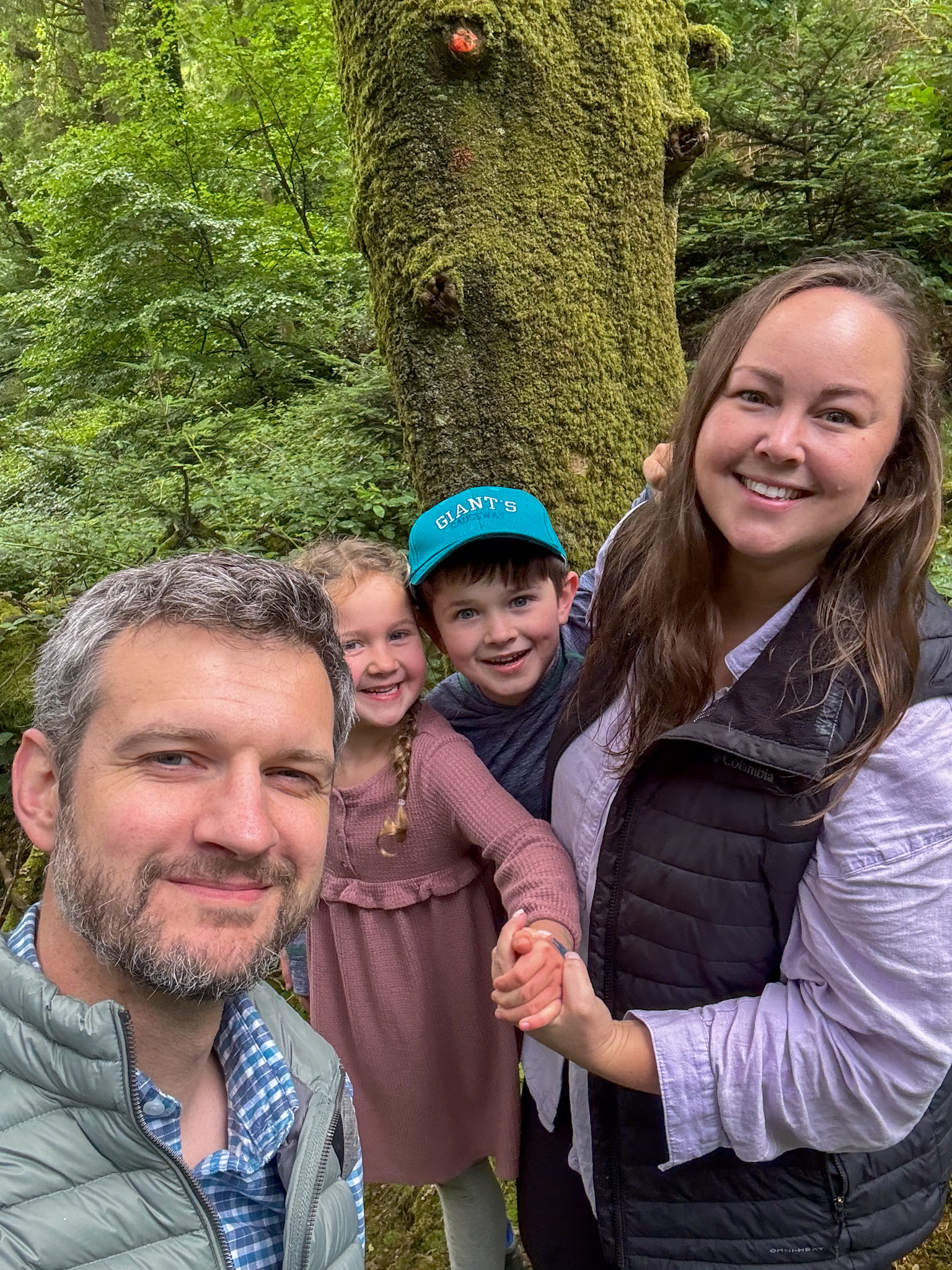 A smiling family of four poses for a selfie in a lush, green forest in Ireland. The father stands in front, with the mother and two children behind him, all holding hands and beaming at the camera. Towering trees covered in moss create a magical, peaceful backdrop for this joyful outdoor moment.
