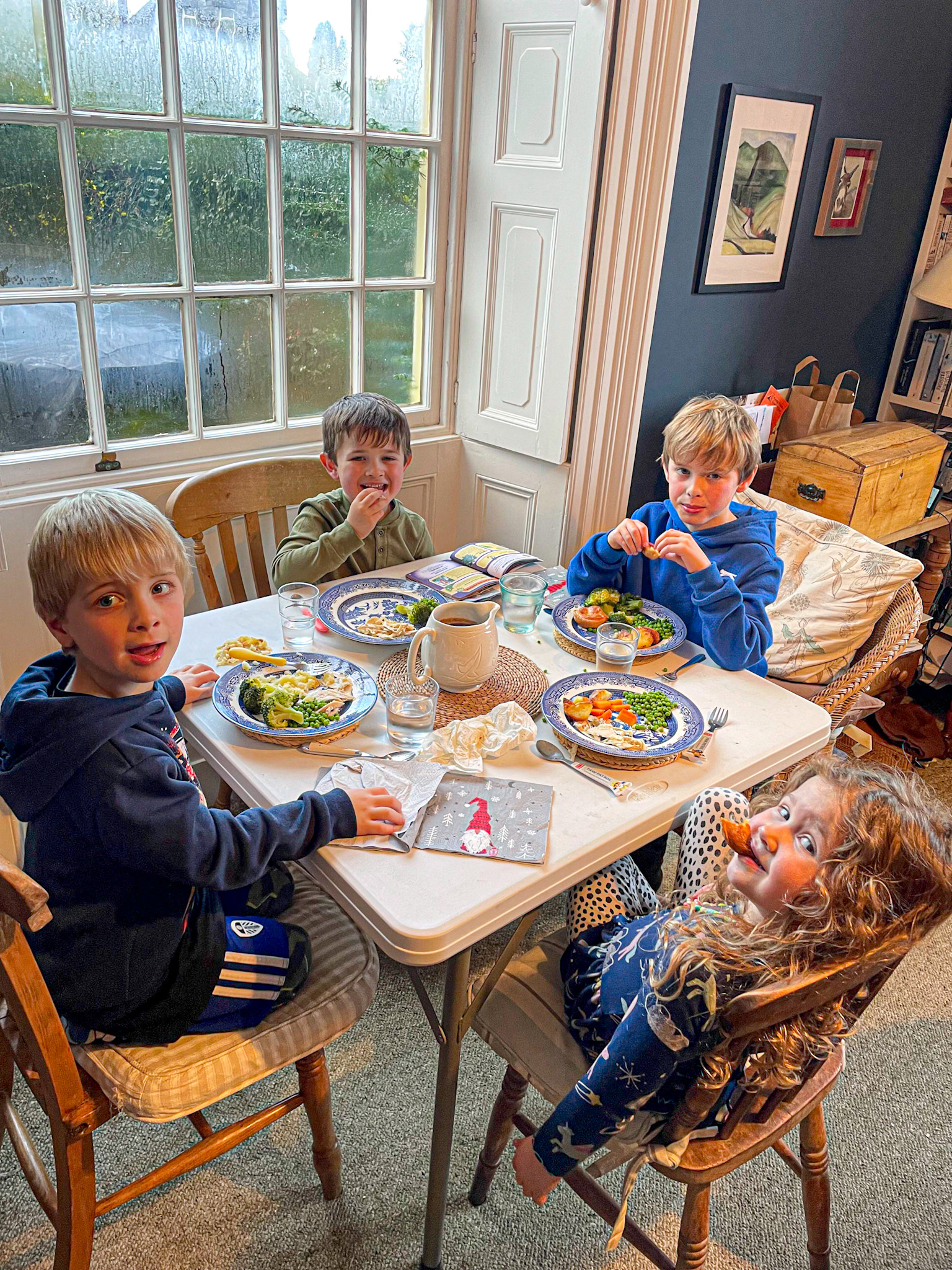 Four young children sit around a cozy dining table enjoying a meal together near a large window with condensation. The kids are smiling and engaged, with plates of vegetables, meat, and potatoes in front of them. The warm, lived-in room has a dark accent wall, framed art, and a mix of wooden chairs, creating a charming, homey atmosphere.