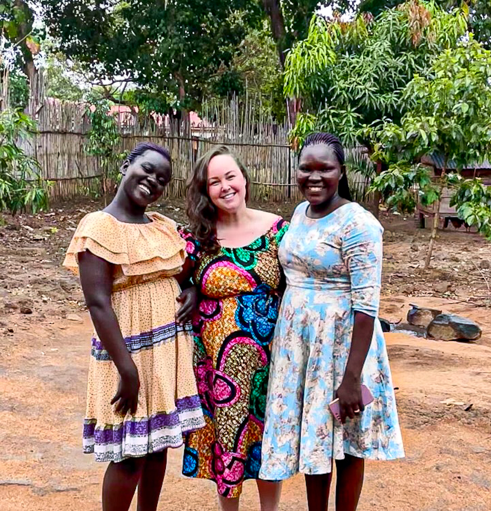 Three women standing close together and smiling, wearing colorful dresses. They are outdoors in a village setting with trees, a bamboo fence, and reddish soil in the background.