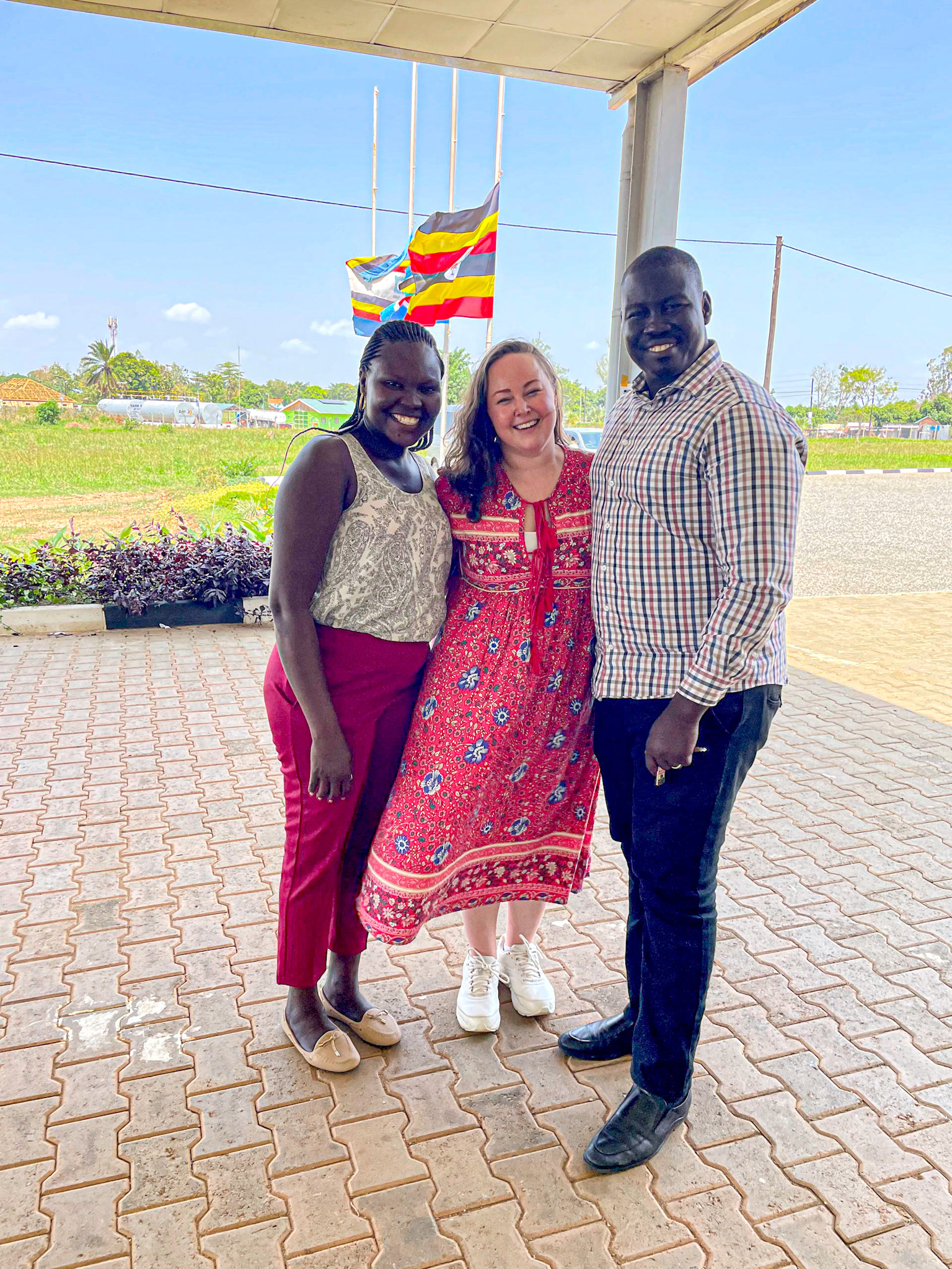 Three people stand smiling together under a shaded area, posing for a photo in front of a bright blue sky with several Ugandan flags waving in the background. The woman in the center wears a red floral dress and white sneakers, while the woman on the left wears red pants and a sleeveless top, and the man on the right wears dark trousers and a checkered shirt. The mood is joyful and relaxed, and the setting appears to be outside a community or government building in Uganda.
