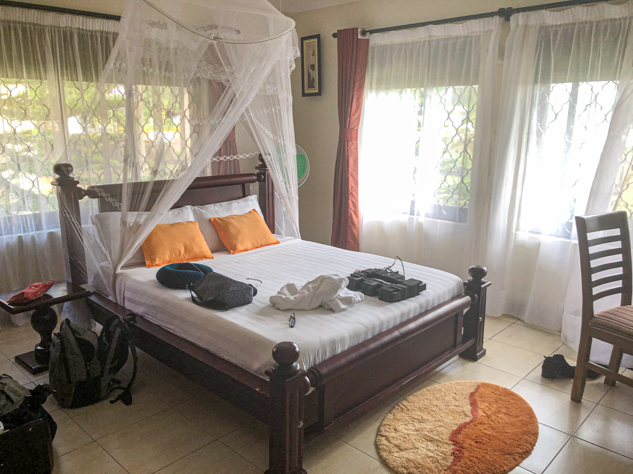 A cozy guesthouse bedroom in Uganda featuring a wooden bed with crisp white linens, bright orange accent pillows, and a white mosquito net draped elegantly overhead. Natural light filters through sheer curtains covering the windows, and travel gear is scattered around the room, suggesting recent arrival. The space feels clean, airy, and welcoming, ideal for a comfortable stay.