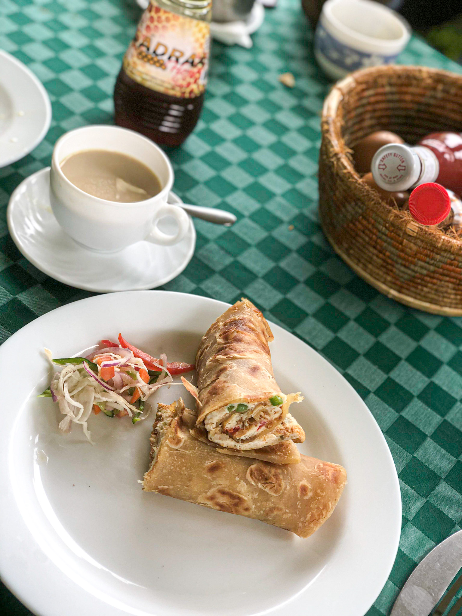 A delicious Ugandan breakfast is served on a white plate featuring a Rolex—an omelette rolled in a chapati—accompanied by a small side of fresh cabbage slaw. The plate sits on a green checkered tablecloth alongside a cup of tea or coffee, a bottle of honey labeled "ADRAX," and a woven basket holding condiments. The setting suggests a cozy, outdoor café or guesthouse meal.