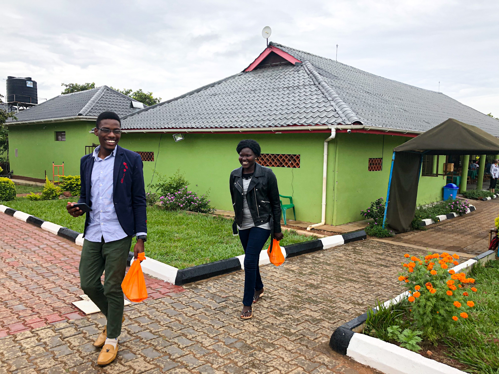 Two smiling people walk along a paved path beside a green building with a grey tiled roof in Uganda. They’re casually dressed and each carry a small orange bag. The area is well-kept with flower beds, a water tank in the background, and a covered walkway. The mood is cheerful and relaxed, set against a cloudy sky.