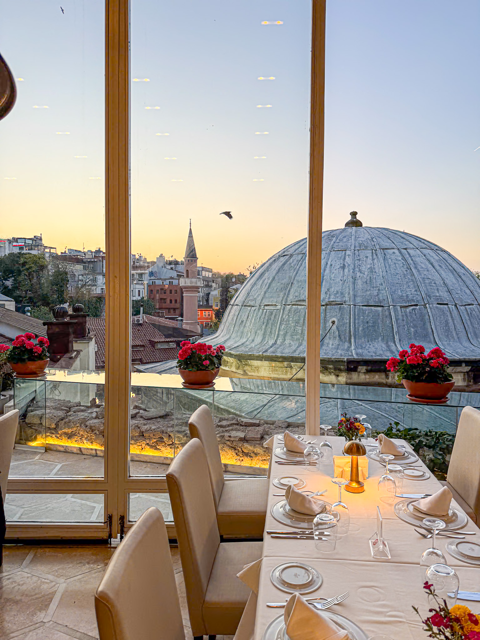 Table set for dinner at a rooftop restaurant in Istanbul with cream-colored linens, glassware, and flowers, overlooking domed architecture and city rooftops at sunset with a bird flying through the sky.