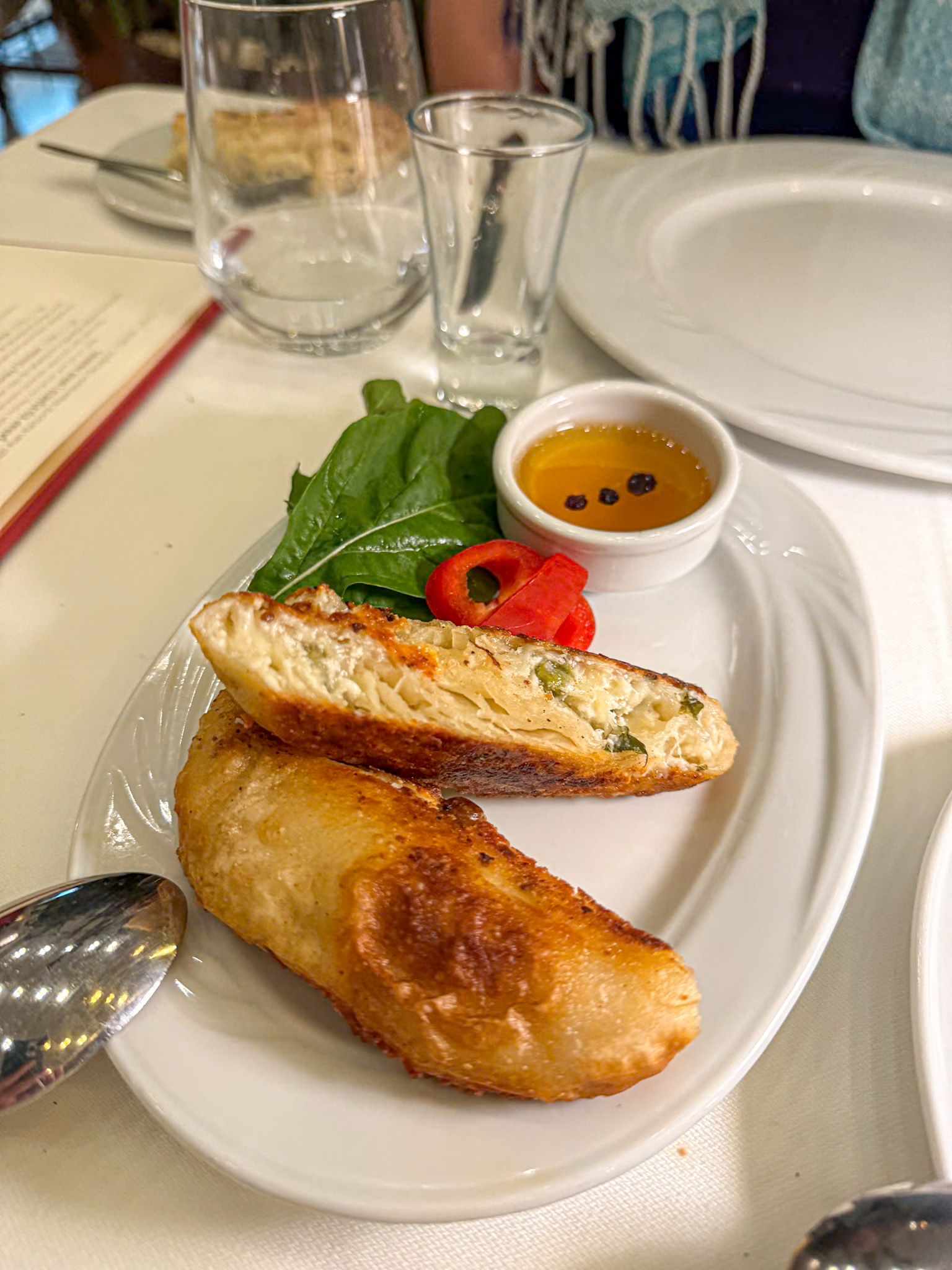 Close-up of a plate with two golden-brown fried pastries filled with cheese and herbs, served with fresh greens, sliced red peppers, and a small dish of dipping sauce, set on a white tablecloth.