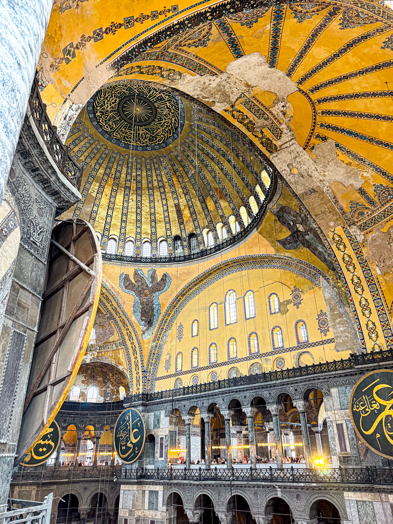Interior view of the Hagia Sophia in Istanbul showcasing its grand domed ceiling, intricate Arabic calligraphy, ancient Christian mosaics, and rows of arched windows and columns.