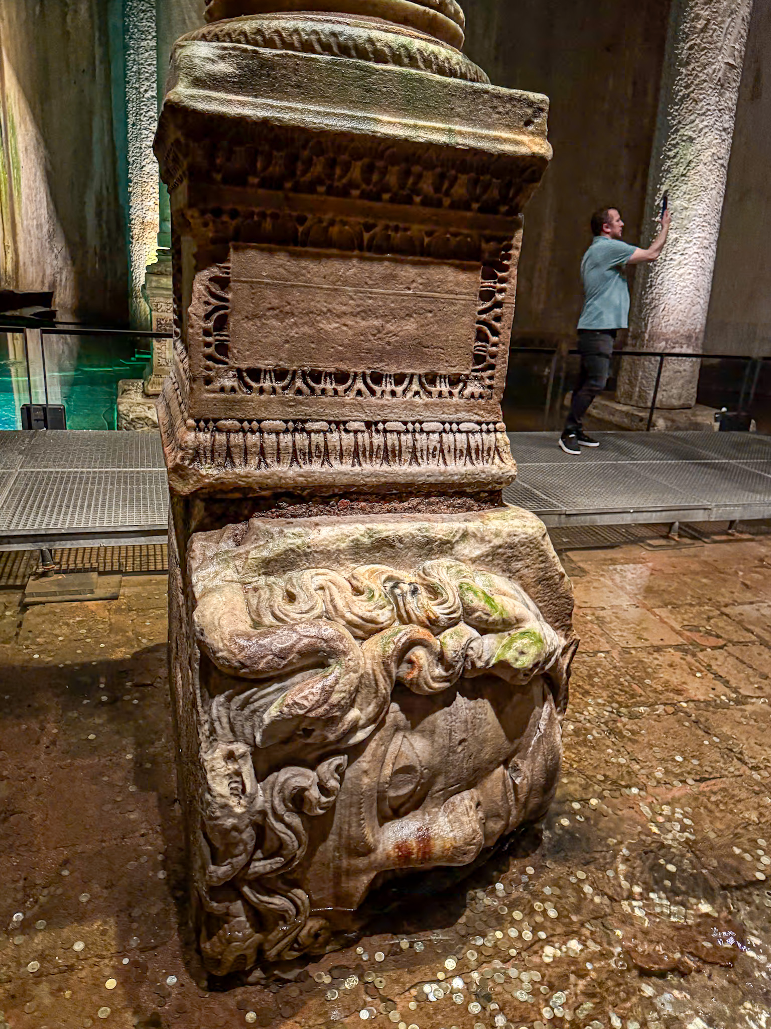 Upside-down Medusa head used as a column base in the Basilica Cistern in Istanbul, partially submerged in water with scattered coins, and a visitor taking a photo in the background.