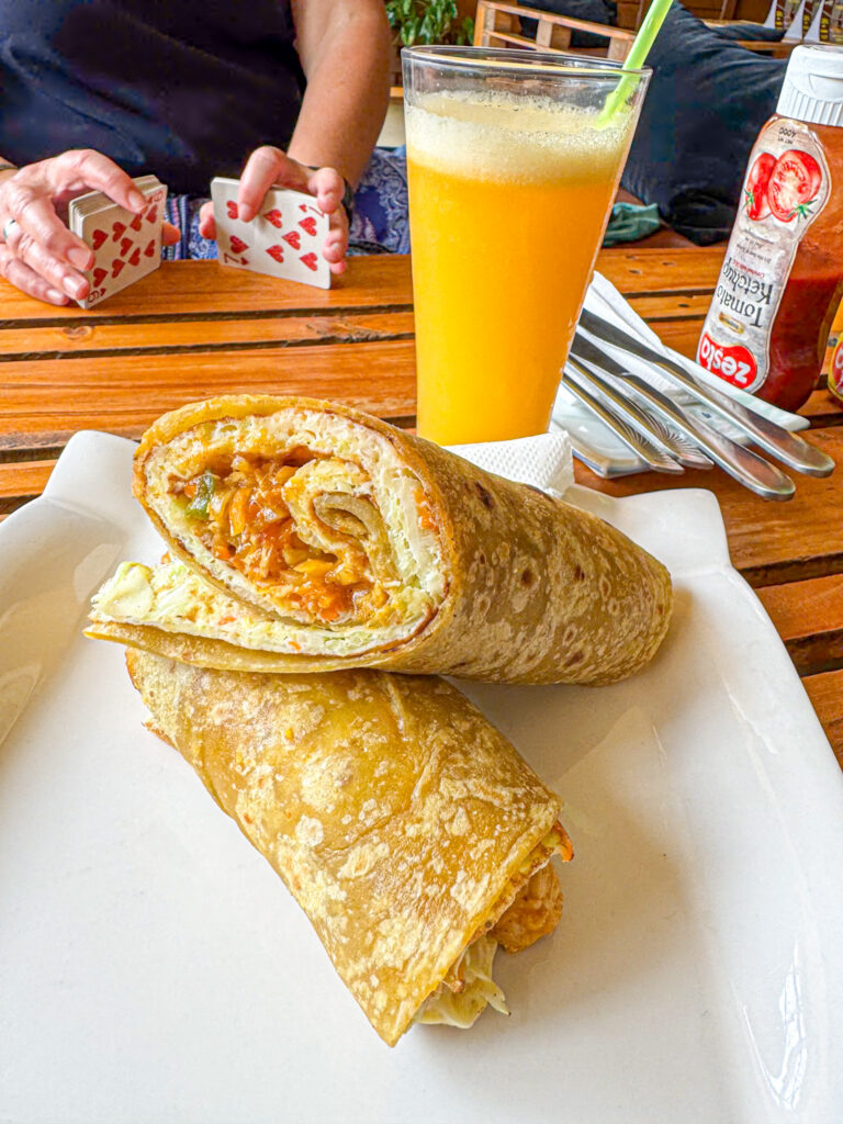 A close up of a Uganda Rolex with eggs and vegetables on a wood table next to a glass of passionfruit juice, someone is playing cards in the background. 