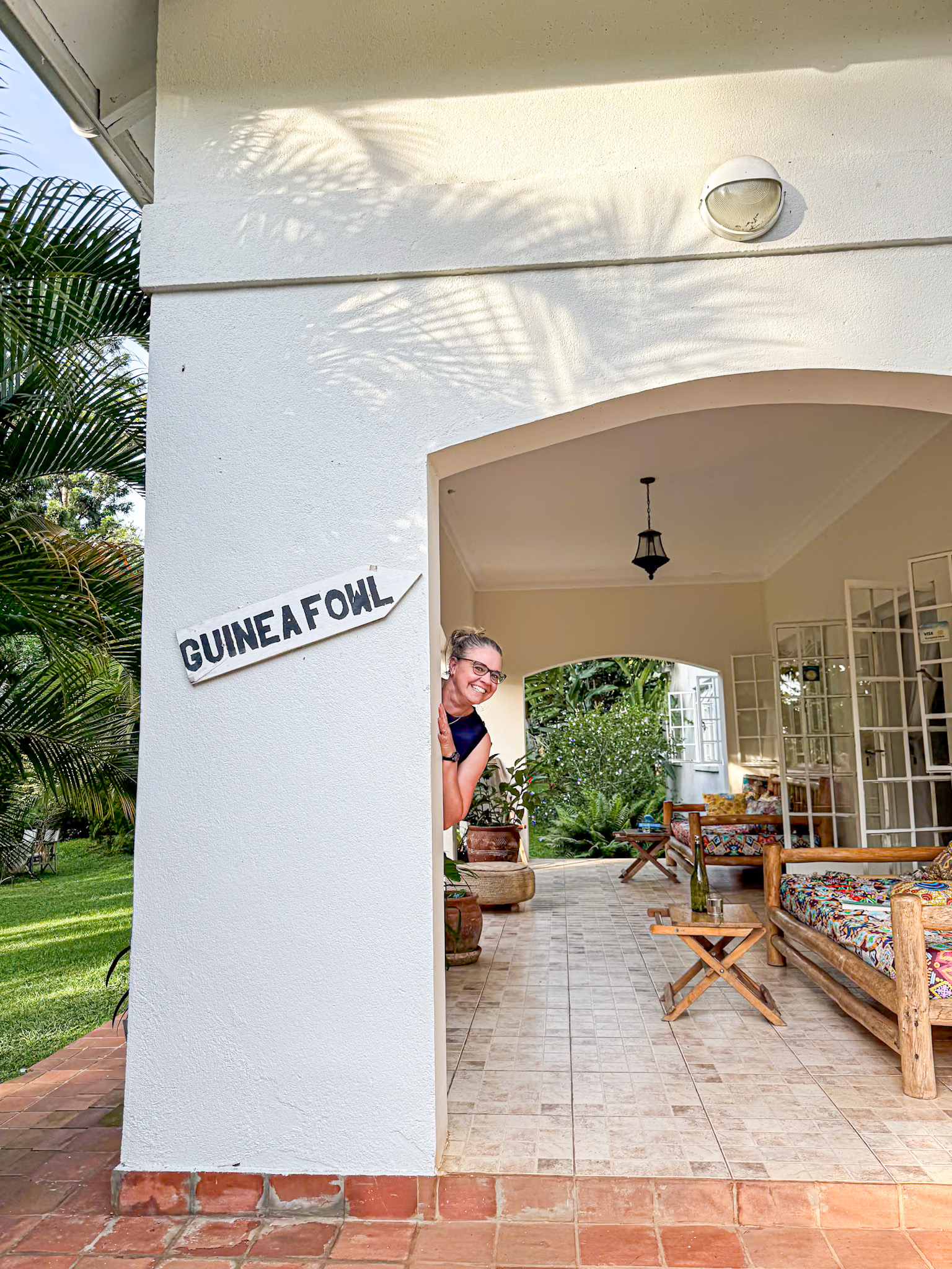 A woman playfully peeks out from behind a white pillar marked with a sign that says "GUINEA FOWL" at a charming guesthouse. The covered veranda features tiled floors, colorful cushioned wooden seating, potted plants, and a peaceful garden view in the background. The scene feels relaxed and welcoming, capturing the cozy atmosphere of a stay in Uganda.