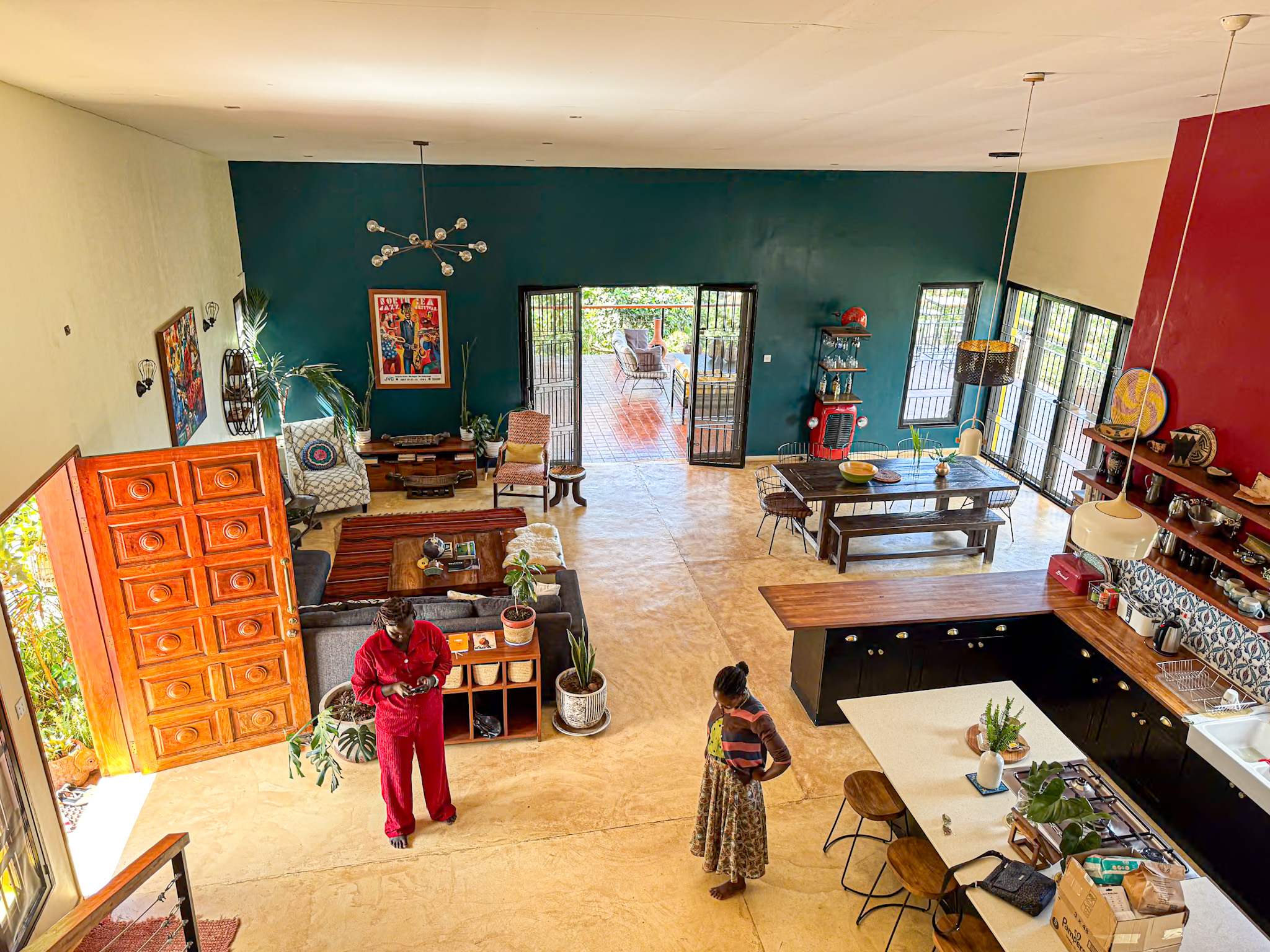 A wide-angle view of a beautifully designed open-concept living space in Uganda, blending modern and local aesthetics. The room features a spacious kitchen with a large island, a long wooden dining table, and a cozy seating area with sofas and colorful artwork. Two people are seen chatting on the polished floor near the entryway, while natural light floods in through large windows and doors that open to a shaded outdoor patio. The décor includes plants, woven baskets, and Afrocentric wall art, creating a warm and inviting atmosphere.