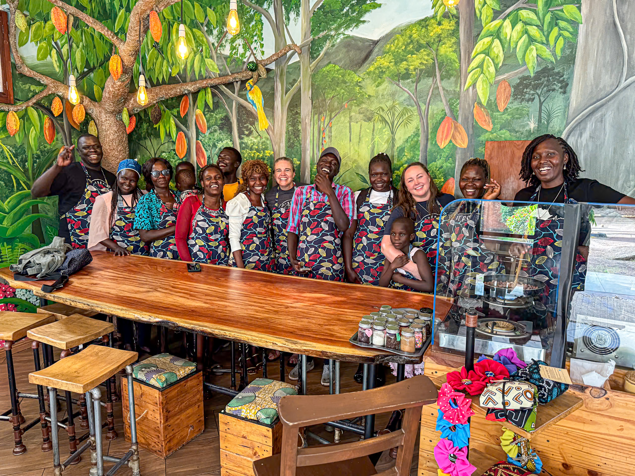 A joyful group of people visiting Uganda, including women, men, and children, stand behind a wooden table inside a vibrant café or classroom decorated with a colorful cocoa tree mural. Everyone is wearing matching aprons with a leaf print and smiling warmly at the camera. The setting includes hanging lightbulbs, wooden stools, jars of spices, and a small machine behind a clear screen, suggesting a hands-on cooking or training workshop. The overall atmosphere is welcoming and full of community spirit.