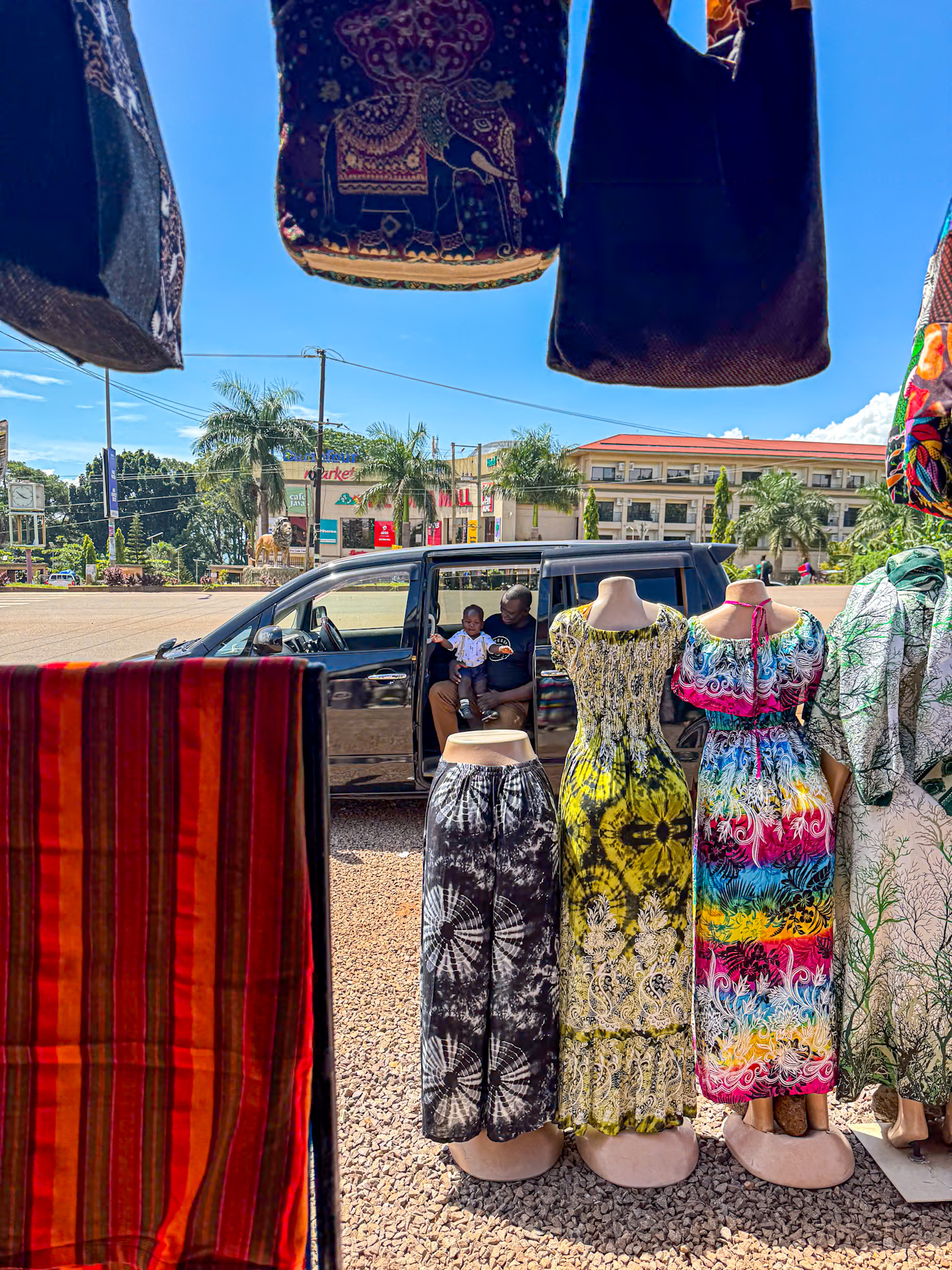 A colorful roadside market scene in Uganda, with vibrant dresses and skirts displayed on mannequins in the foreground. Traditional fabric bags and cloths hang overhead, framing the image. In the background, two people sit in an open van door, smiling and talking, with palm trees and commercial buildings visible across the street under a bright blue sky. The setting captures the lively and sunny atmosphere of a typical shopping stop in town.