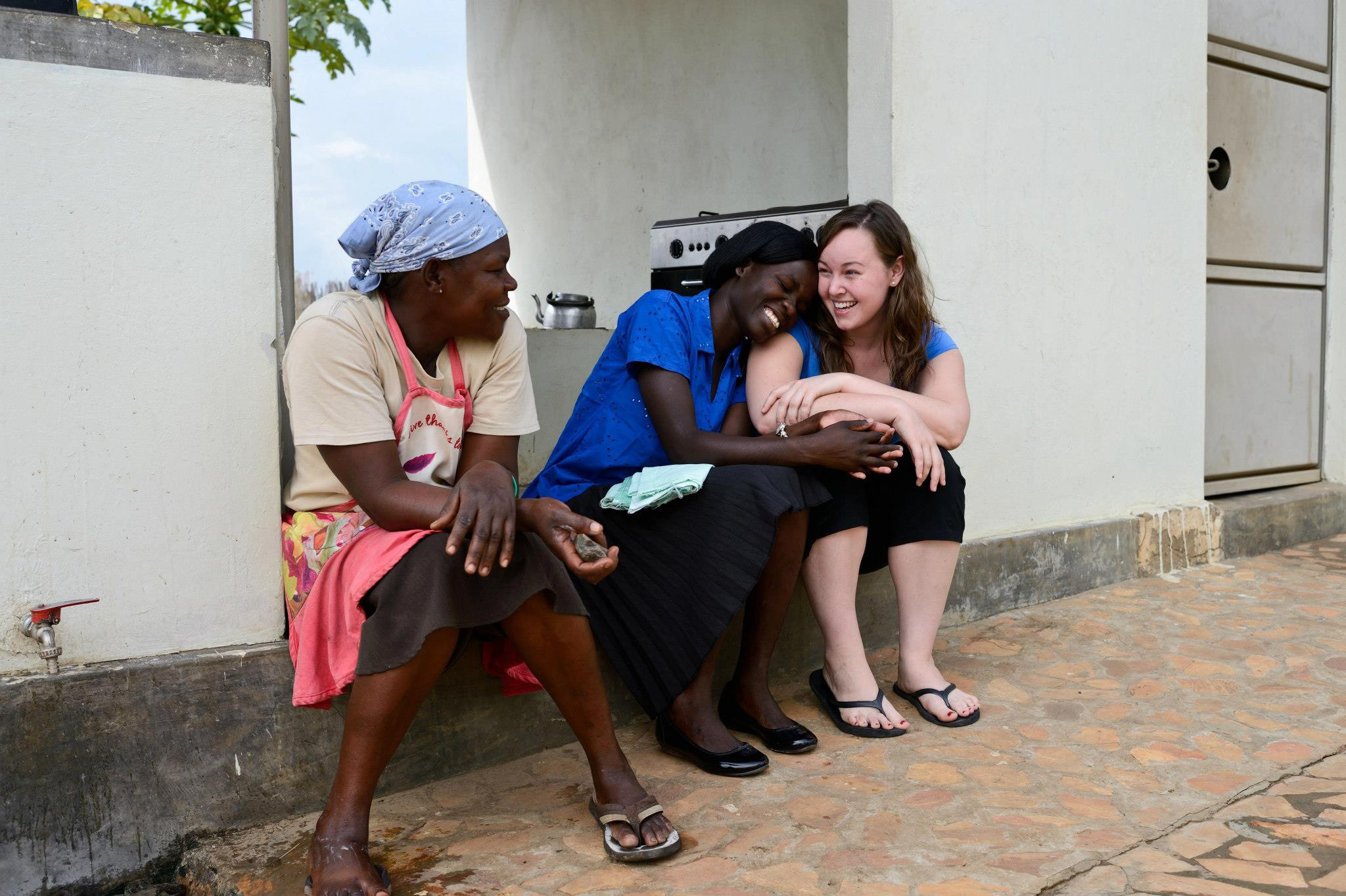 Three women, one is Elizabeth Jordan-Flight, sit together on a stone patio outside a building, laughing and enjoying each other's company. One woman wears an apron and headscarf, another is dressed in a blue blouse and black skirt, and the third is in a blue top and black pants. Their expressions and body language show warmth, joy, and friendship. A kettle and stove can be seen in the background.