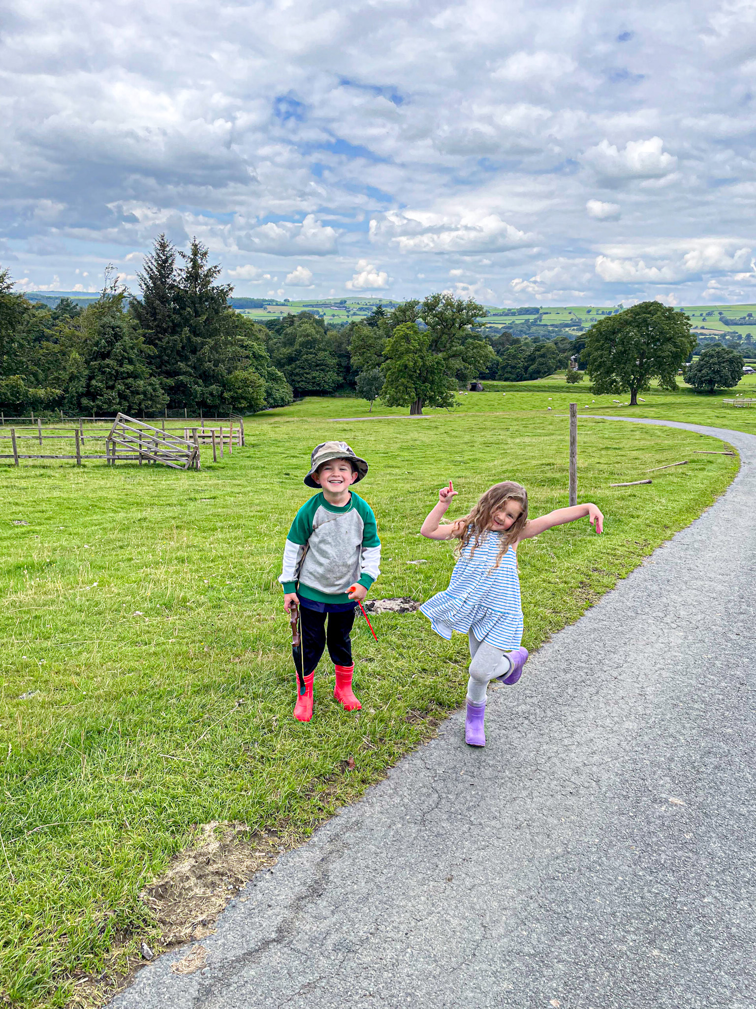 Two young children play and smile on a country road surrounded by lush green fields and rolling hills. The boy wears a bucket hat, red rain boots, and a green and gray sweatshirt, while the girl twirls in a striped dress and purple boots. The sky is filled with fluffy clouds, and the scene is full of vibrant, rural charm.