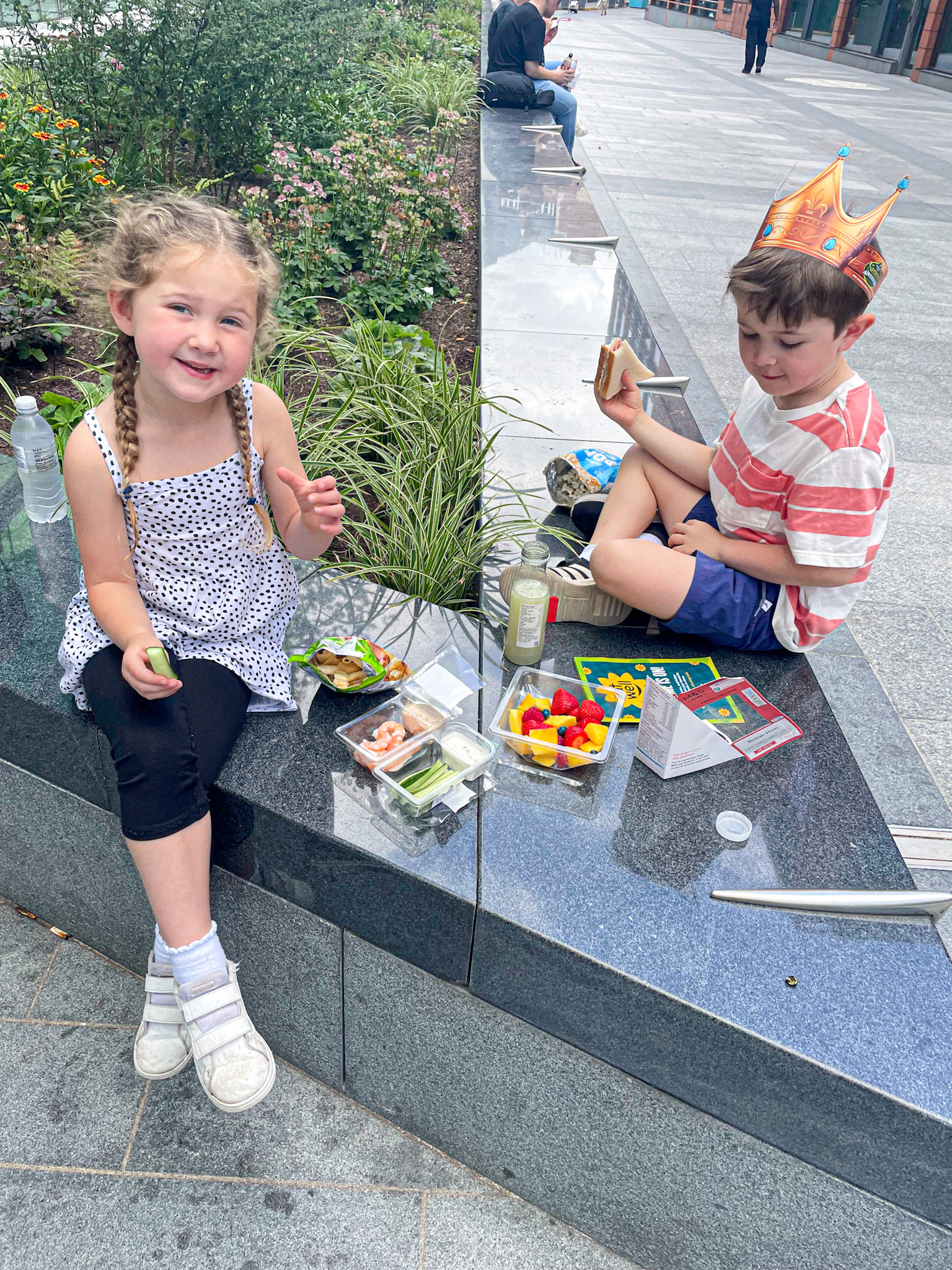 Two young children enjoy a casual picnic on a granite bench in London. The girl, smiling with braids and wearing a polka dot top, holds a snack while the boy, wearing a crown and striped shirt, eats a sandwich. Between them are fruit, veggie packs, and drinks, with flowers and greenery in the background. 