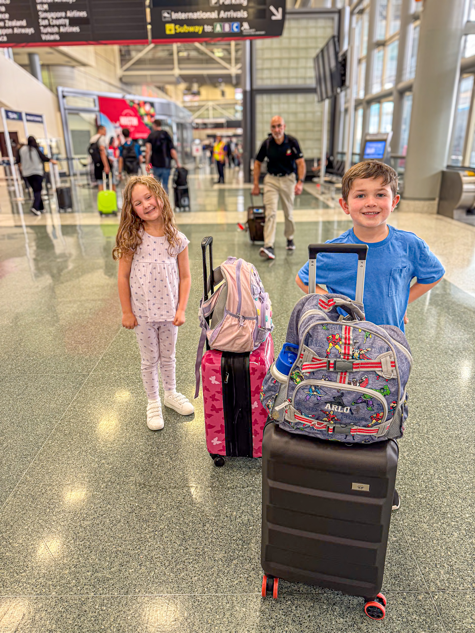 Two smiling children stand proudly with their rolling suitcases in an airport terminal under a sign for International Arrivals. The girl wears light pink pajamas with a pattern, and the boy wears a blue T-shirt with his hands behind his back. Their colorful bags are stacked and ready for travel, capturing the excitement of an international adventure.