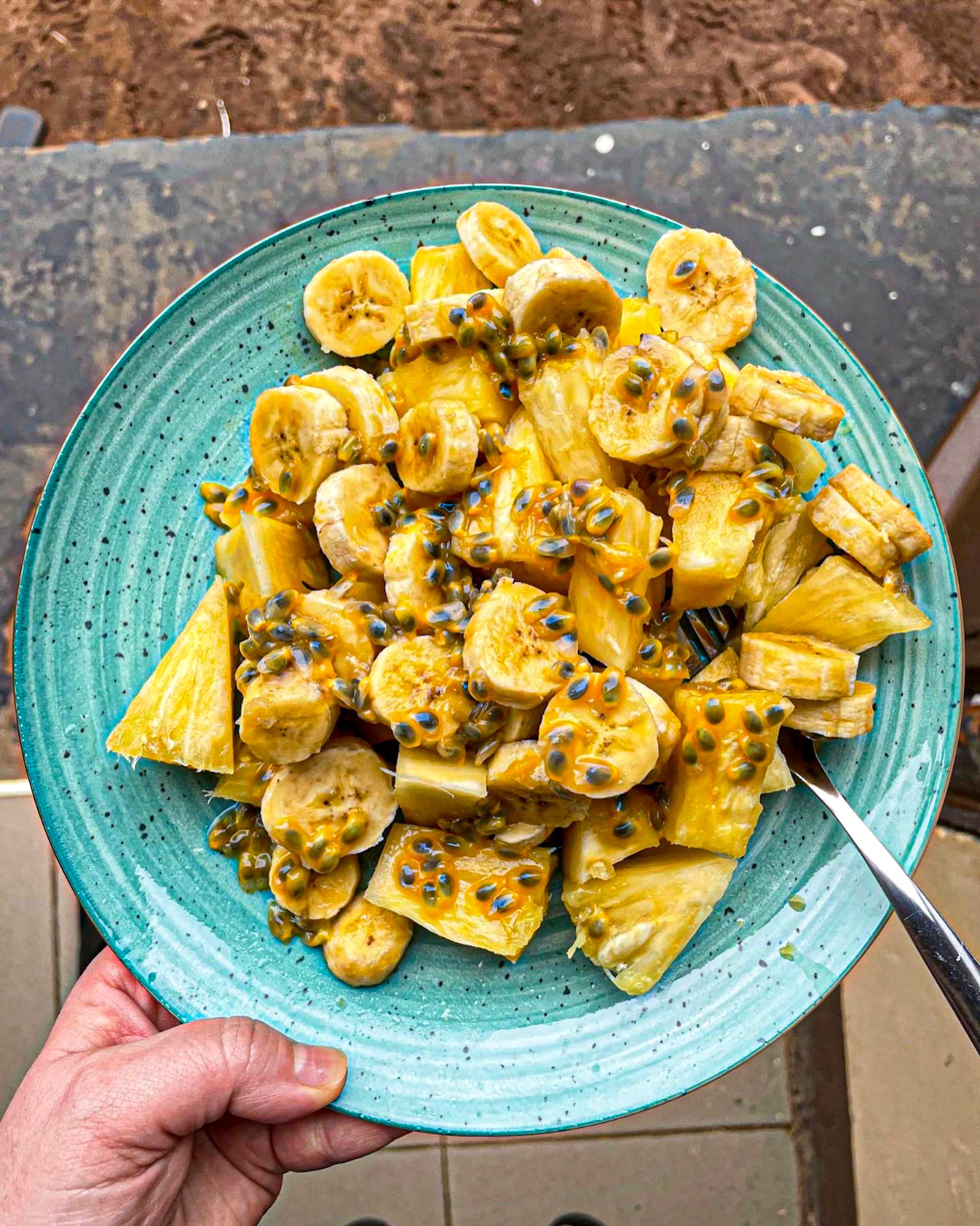 A hand holding a turquoise plate filled with fresh tropical fruit salad made of sliced bananas, pineapple chunks, and passion fruit pulp, with a fork resting on the side.