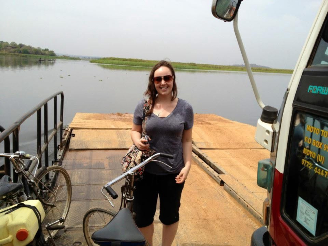 Woman standing on a ferry in East Africa holding a bicycle, smiling at the camera. She is wearing sunglasses, a gray t-shirt, and black shorts. A van and another bicycle are visible beside her. The ferry is on a calm river with grassy banks in the background under a hazy sky.