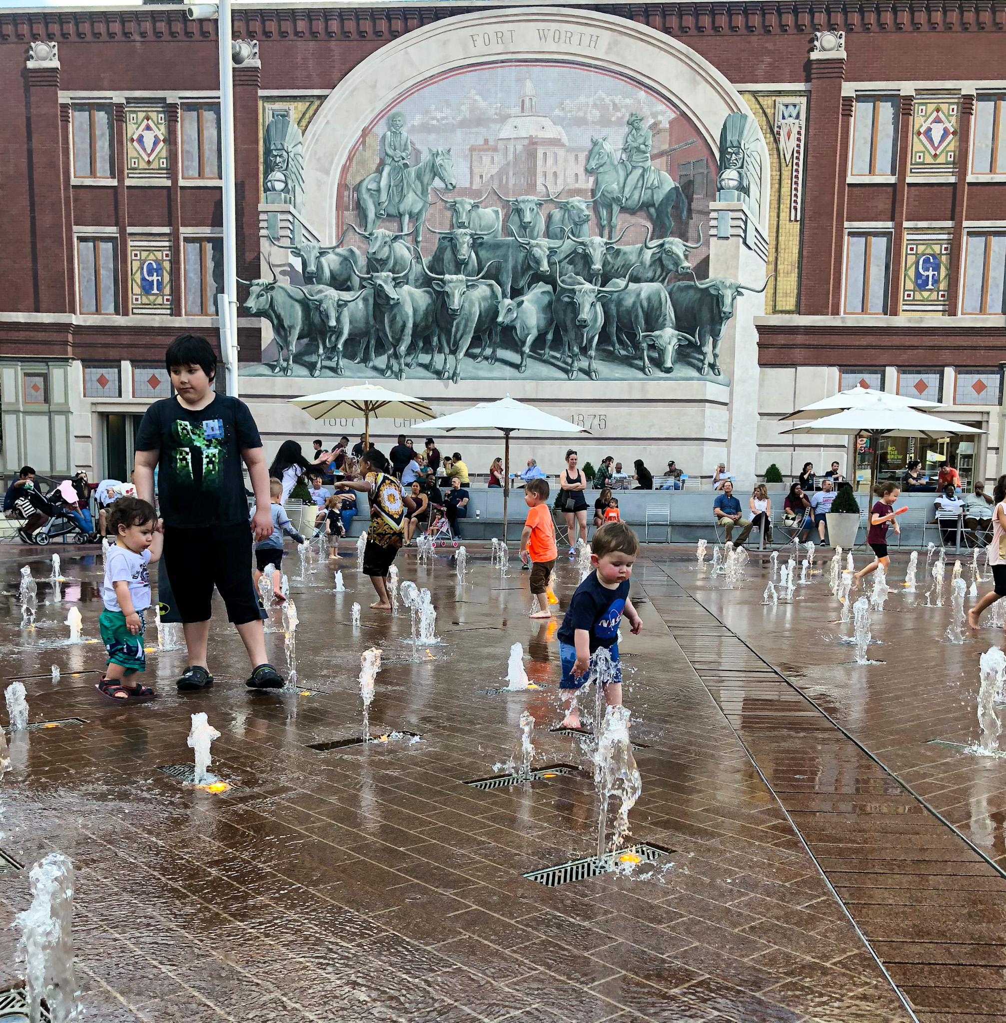 Children play in a splash pad with water jets shooting from the ground in a public plaza, while adults sit and watch in the background. A large mural of cattle and cowboys labeled "Fort Worth" decorates the brick building behind them.