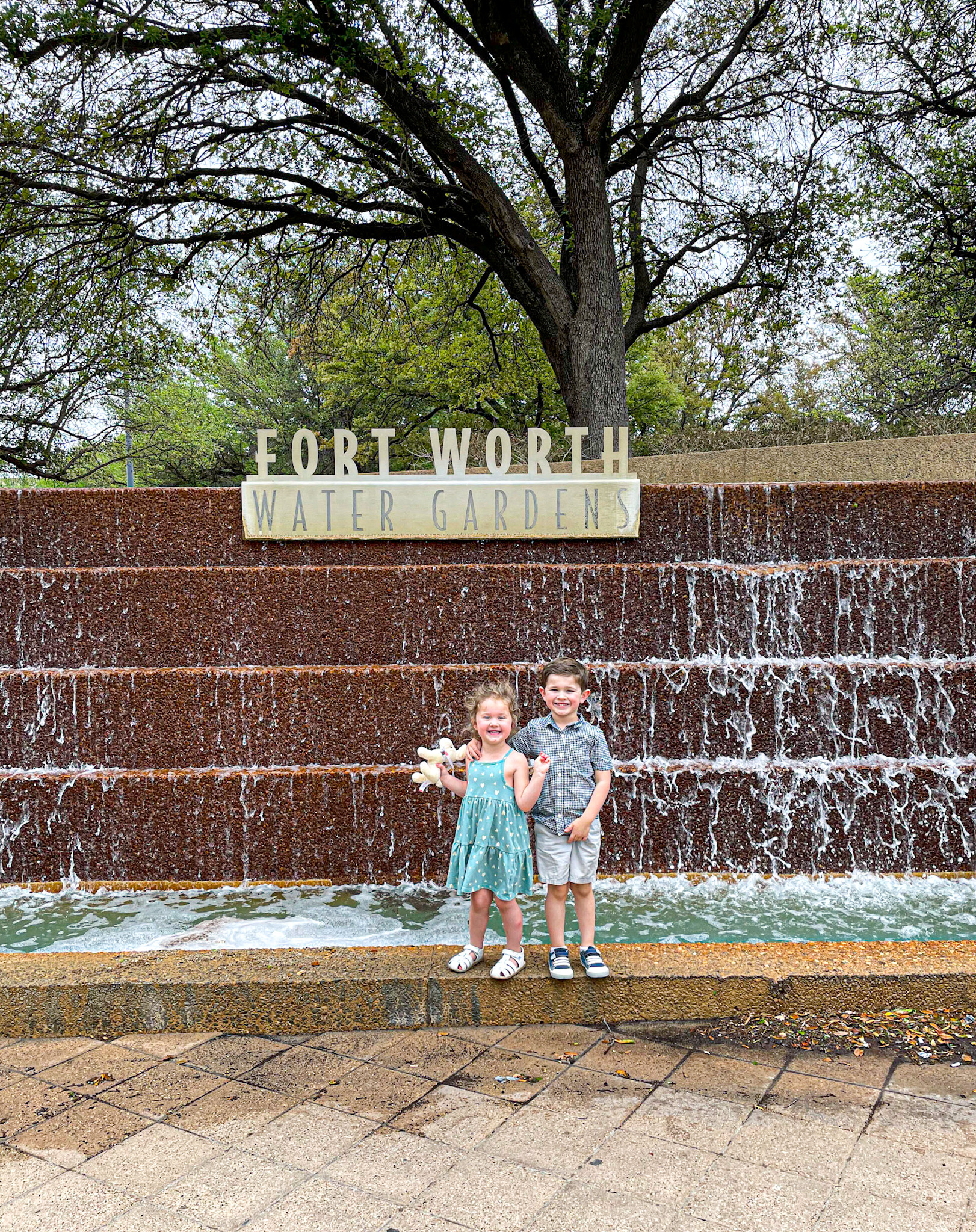 Two young children smile and pose in front of a cascading waterfall feature at the Fort Worth Water Gardens. The girl in a teal dress holds a stuffed animal, while the boy in a gray shirt and shorts stands beside her. Trees with early spring foliage are visible above the “Fort Worth Water Gardens” sign.