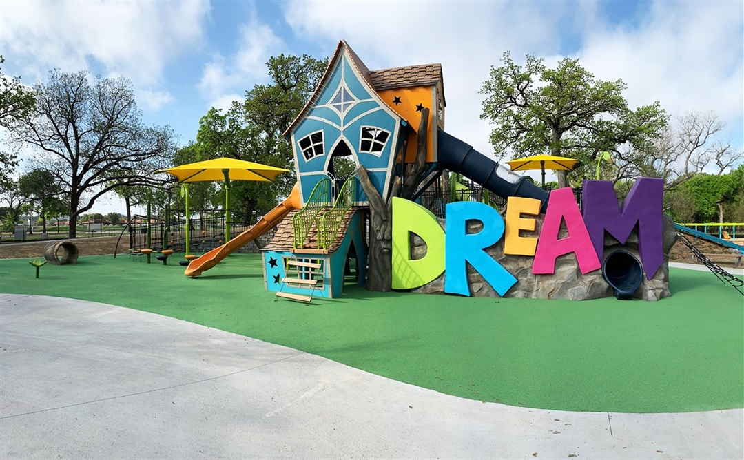 A colorful, whimsical playground features a tilted playhouse structure with slides and climbing features, surrounded by the large word “DREAM” in bright, oversized letters. The play area has a green cushioned surface, yellow shade umbrellas, and is bordered by trees under a partly cloudy sky.
