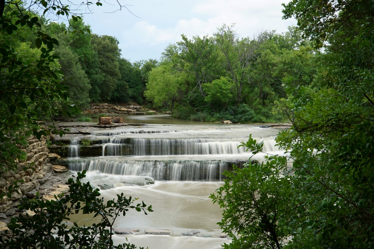 A serene multi-tiered waterfall flows gently over limestone rocks, surrounded by lush green trees and dense foliage. The smooth, cascading water creates a peaceful atmosphere in this natural woodland setting.