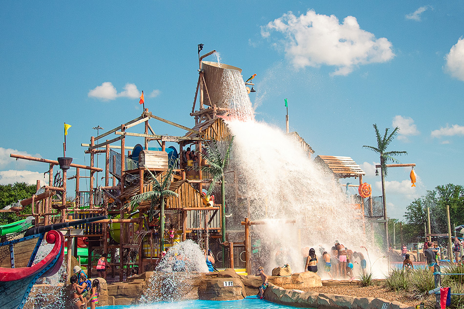 Large bucket dumping water at a busy outdoor water park called Hurricane Harbor in Arlington, Texas on a sunny day, with children playing below.