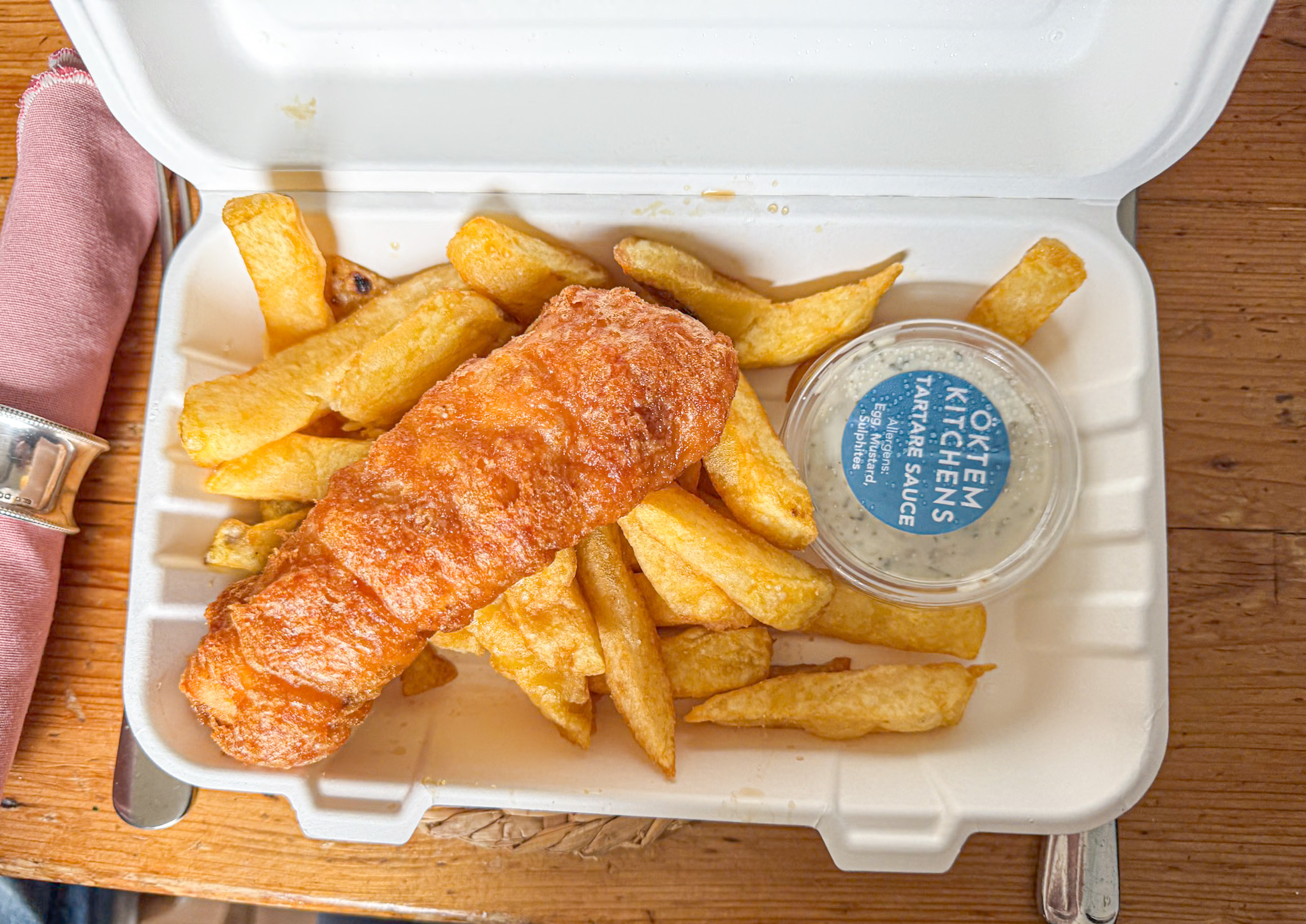 Fish and chips in a takeout box with tartar sauce on a wooden table.