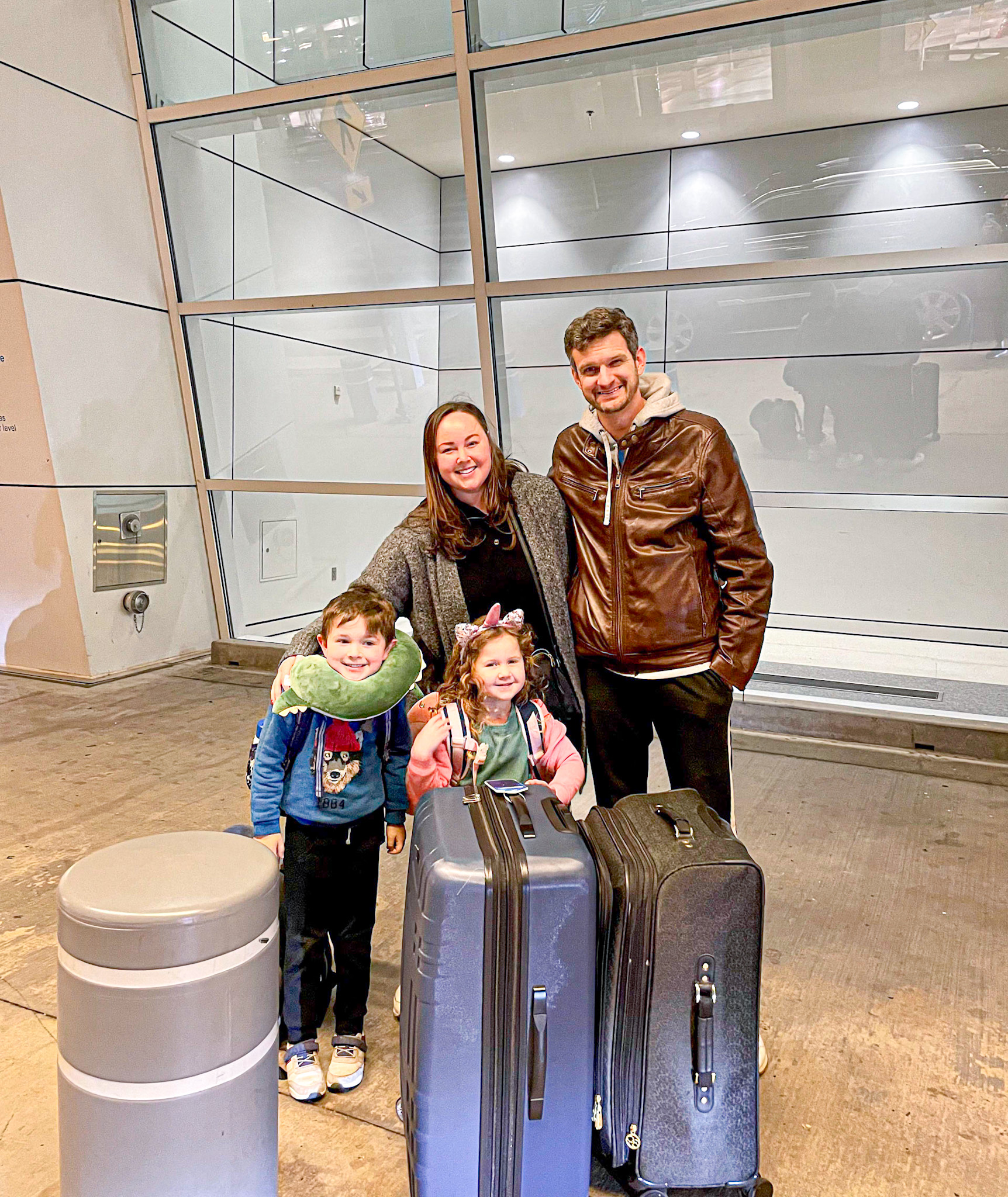 A smiling family of four with luggage stands outside an airport terminal, ready for their trip traveling internationally with kids.