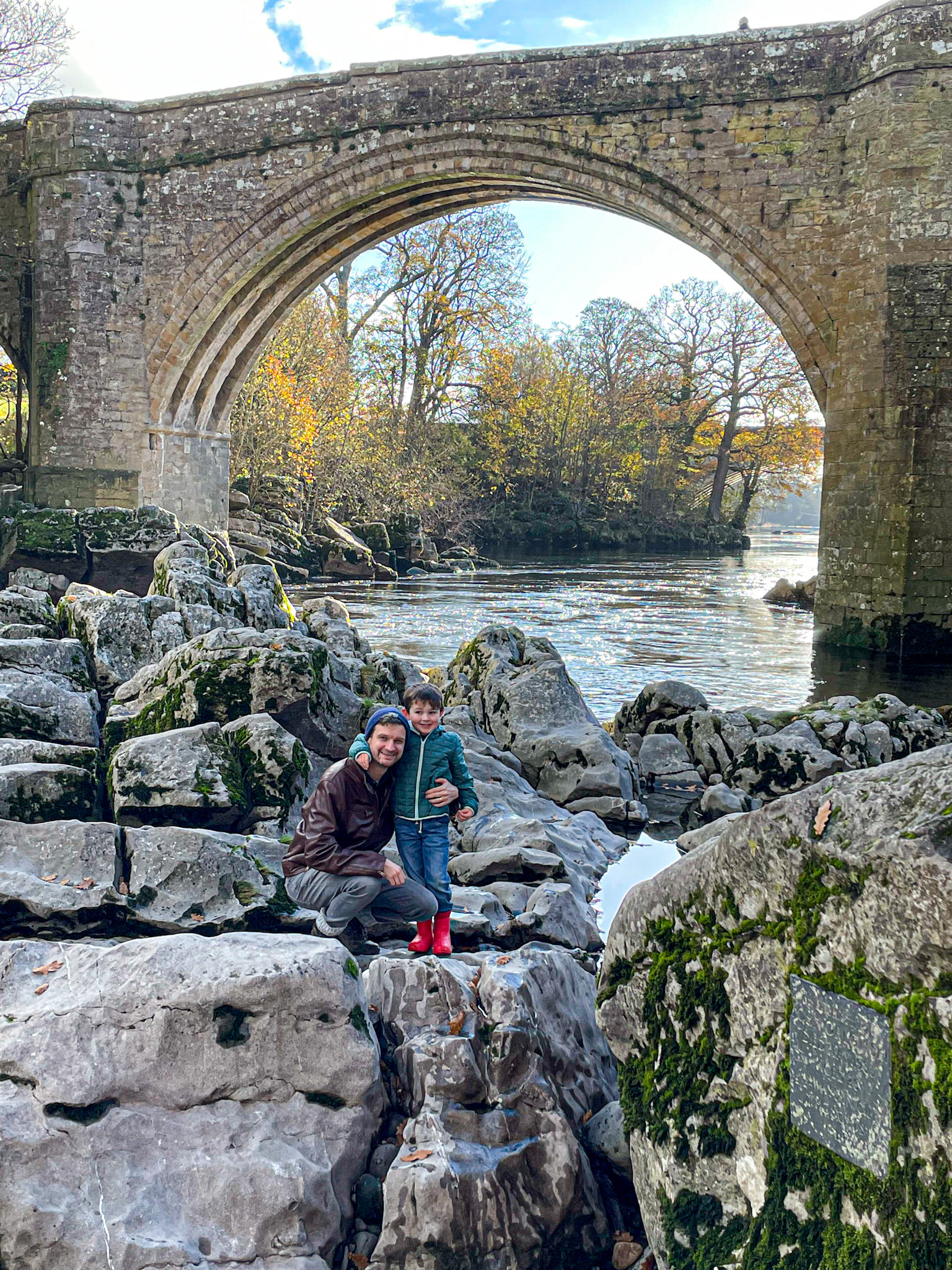 An adult and child stand on rocks by a river under an old stone arch called Devil's bridge surrounded by autumn trees in the English lake district.