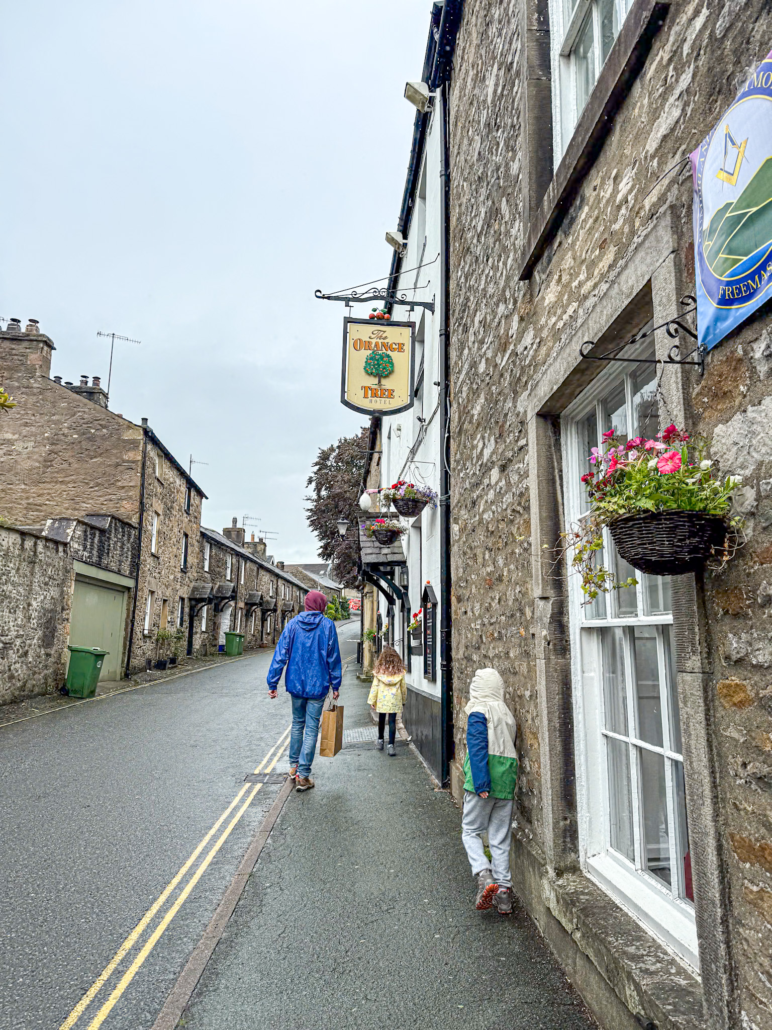 Three people walk down a wet street beside stone buildings with hanging flower baskets and a pub sign.
