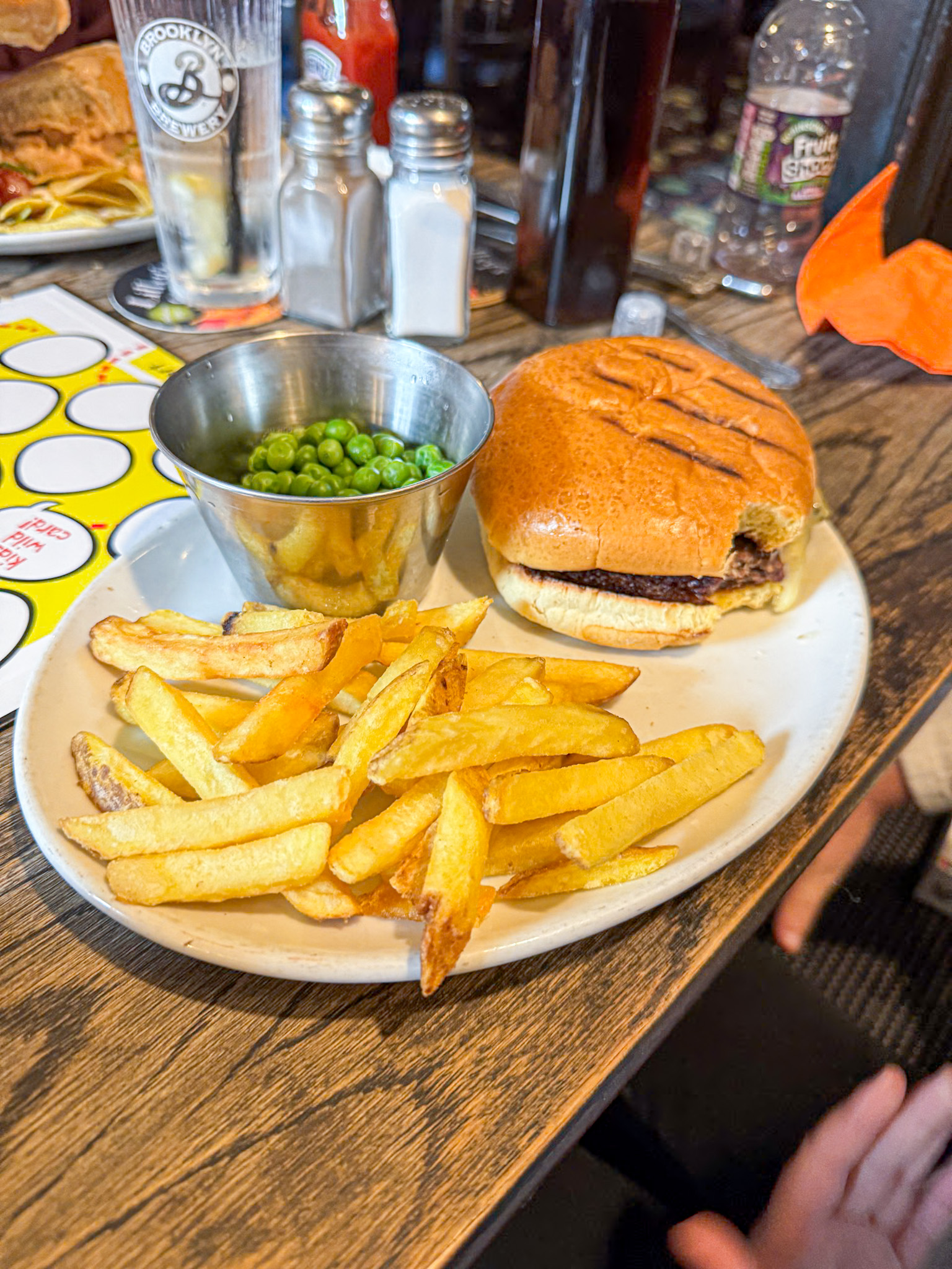 Plate with a burger, fries, and a metal cup of peas on a wooden table in a restaurant setting.