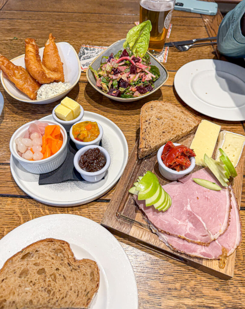 A variety of cold meats, cheeses, bread, salad, and pickles served on a wooden table in a pub setting.