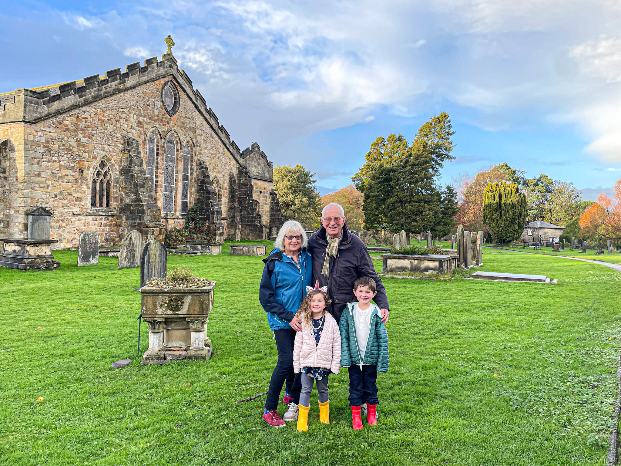 Two adults and two children smiling in front of an St. Mary's Church and gravestones on a grassy field in Kirkby Lonsdale. 