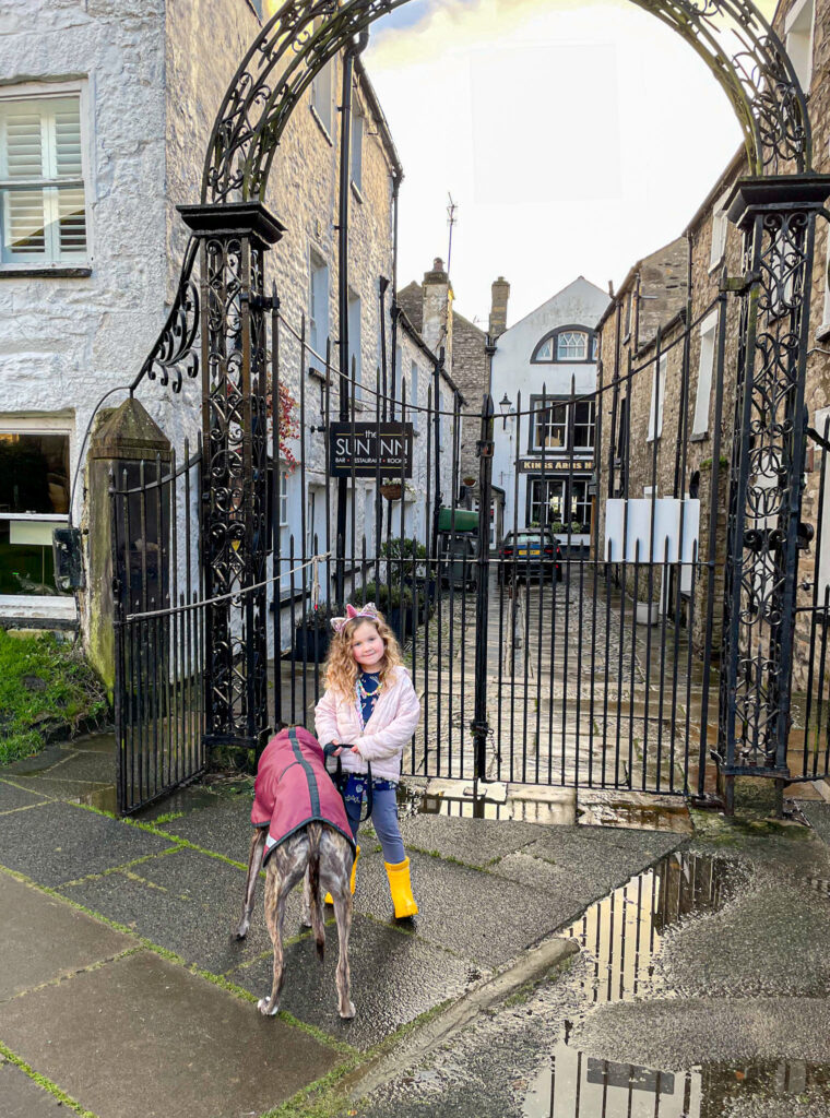 A smiling girl in yellow boots holds a greyhounds leash by a wrought iron gate in a stone-paved alley.
