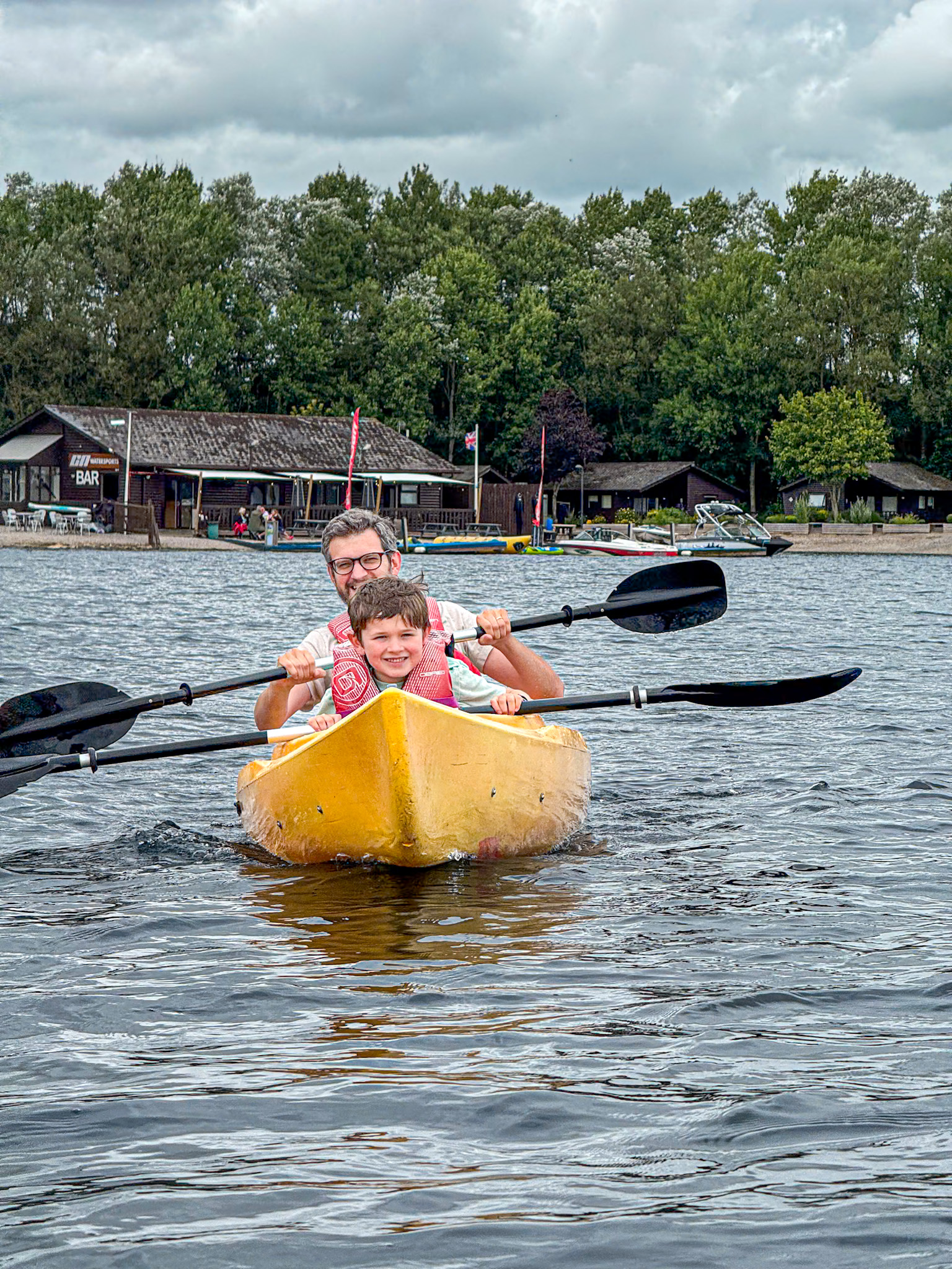 A man and child kayaking on a lake, smiling, with trees and buildings in the background.