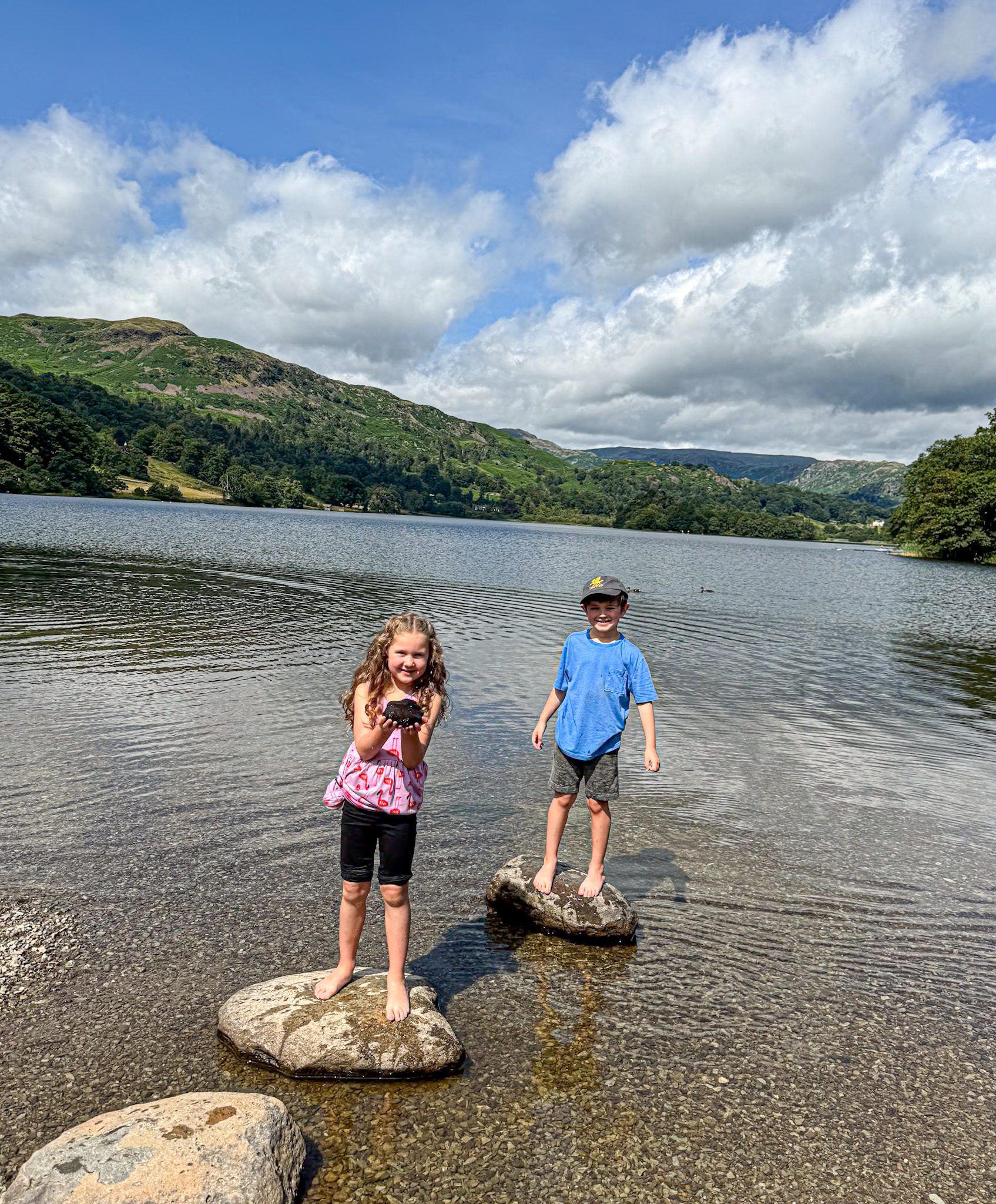 Two children stand barefoot on rocks in a lake with green hills and a cloudy sky in the background.