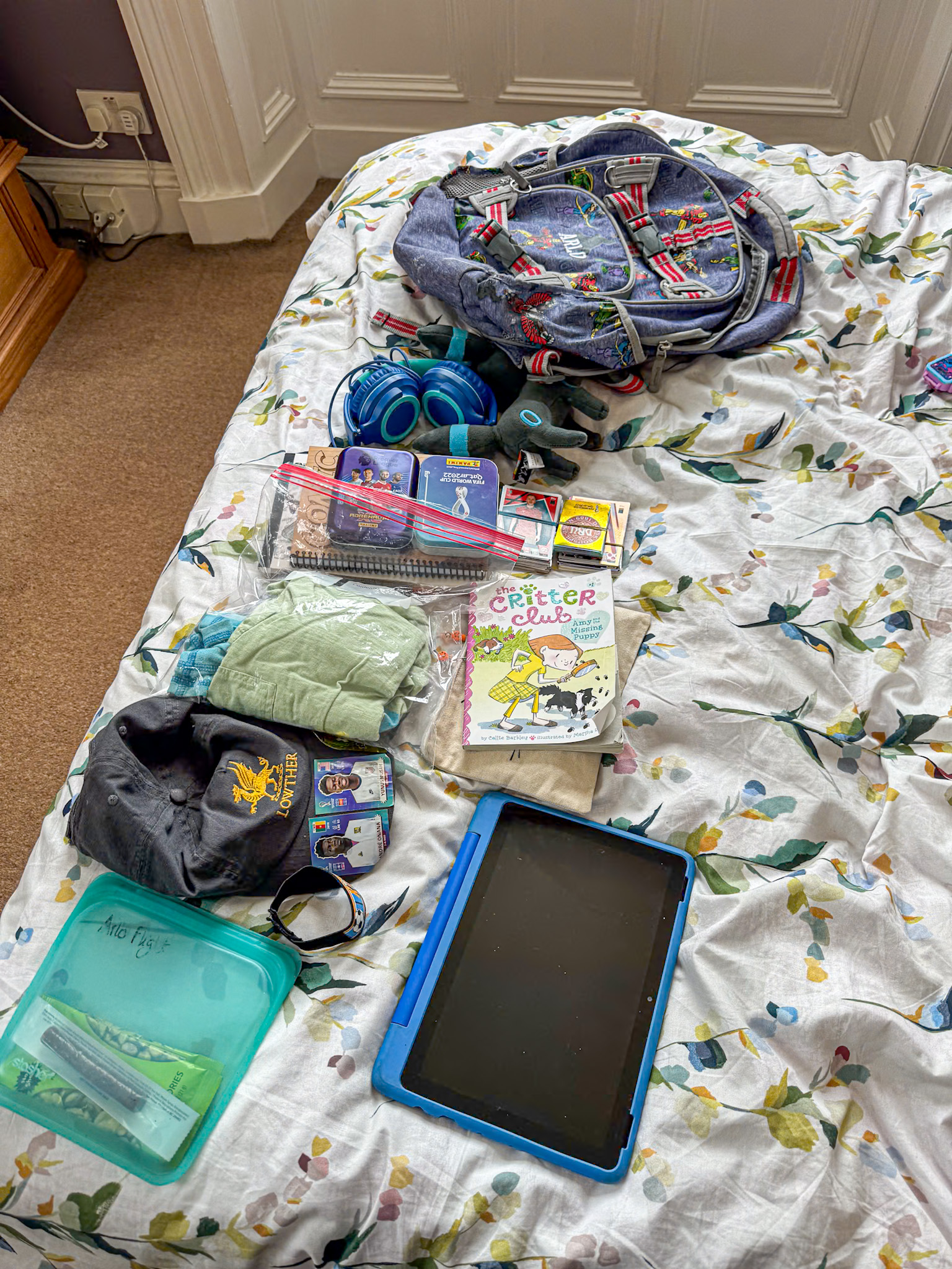A backpack, headphones, clothes, books, a pencil case, and a tablet laid out on a bed with a floral sheet, all you need for traveling internationally with kids. 