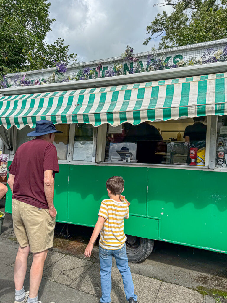 An adult and child stand at a green food truck with a striped awning on a cloudy day.