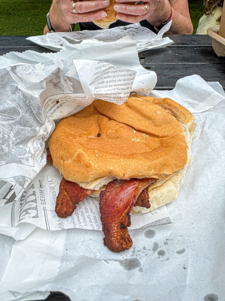 A bacon sandwich on bread sits on newspaper with a person holding food in the background.
