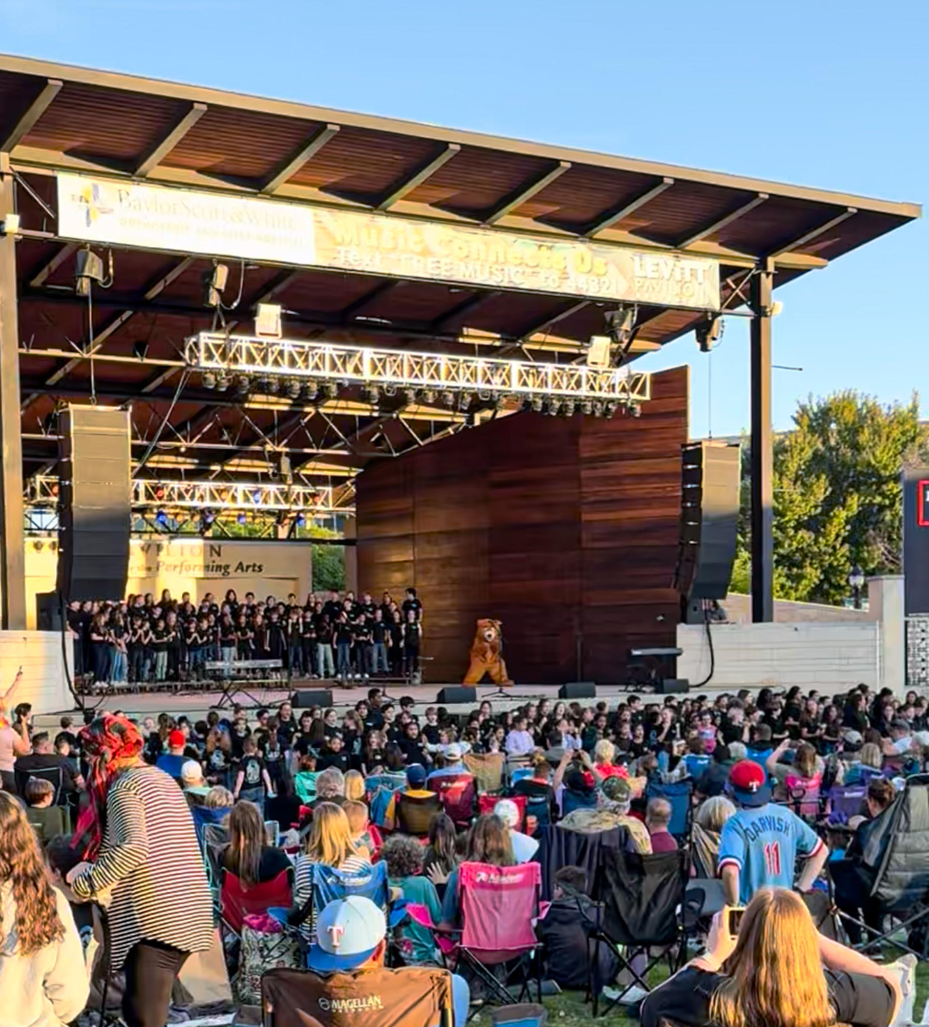 Outdoor concert at Levitt Pavilion in Arlington, Texas with choir and a mascot on stage, large audience seated in lawn chairs on a sunny day.