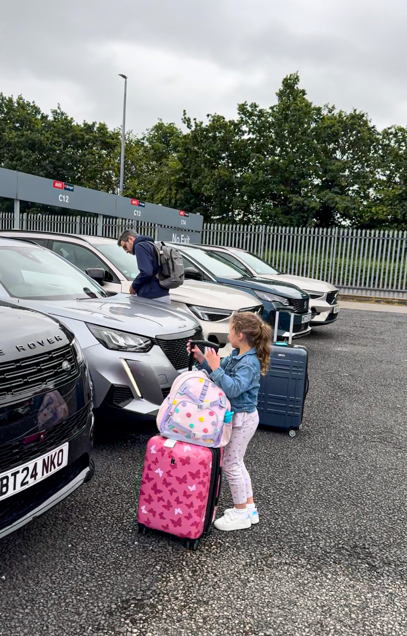A girl with pink luggage stands by cars in a parking lot, while a man loads bags into a silver car.