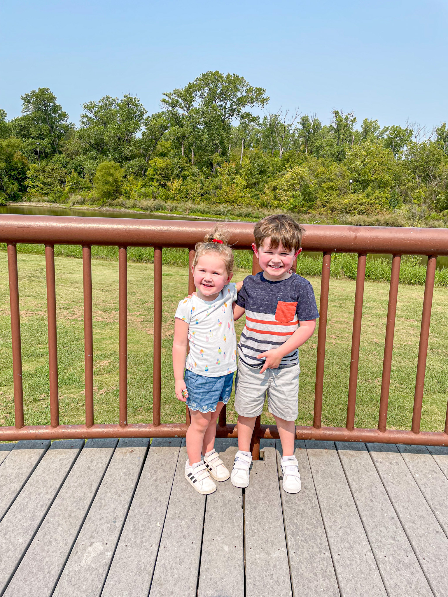 Two young children smiling and standing together on a wooden bridge with greenery in the background at River Legacy Park. 