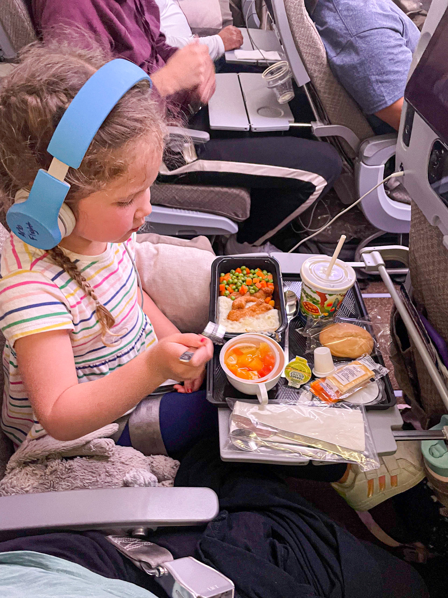 A young girl with headphones eats a meal on an airplane, with food and drink on a tray table.