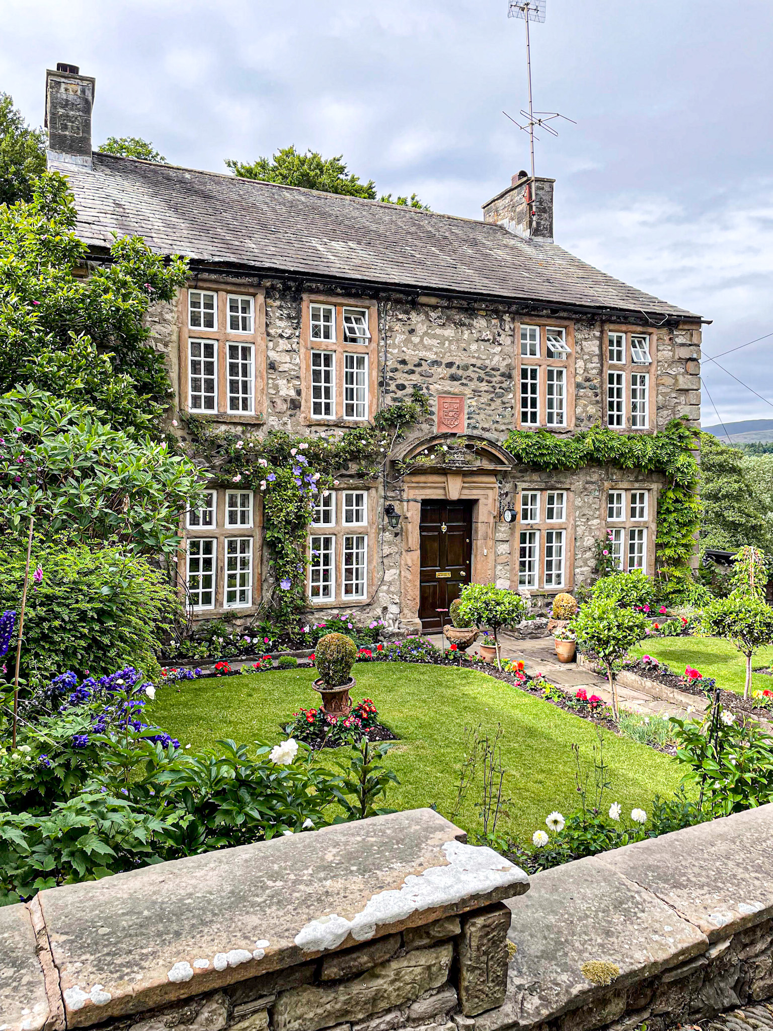 Stone house called the Old Manor in Kirkby Lonsdale with large windows, surrounded by lush gardens and blooming flowers on a sunny day.
