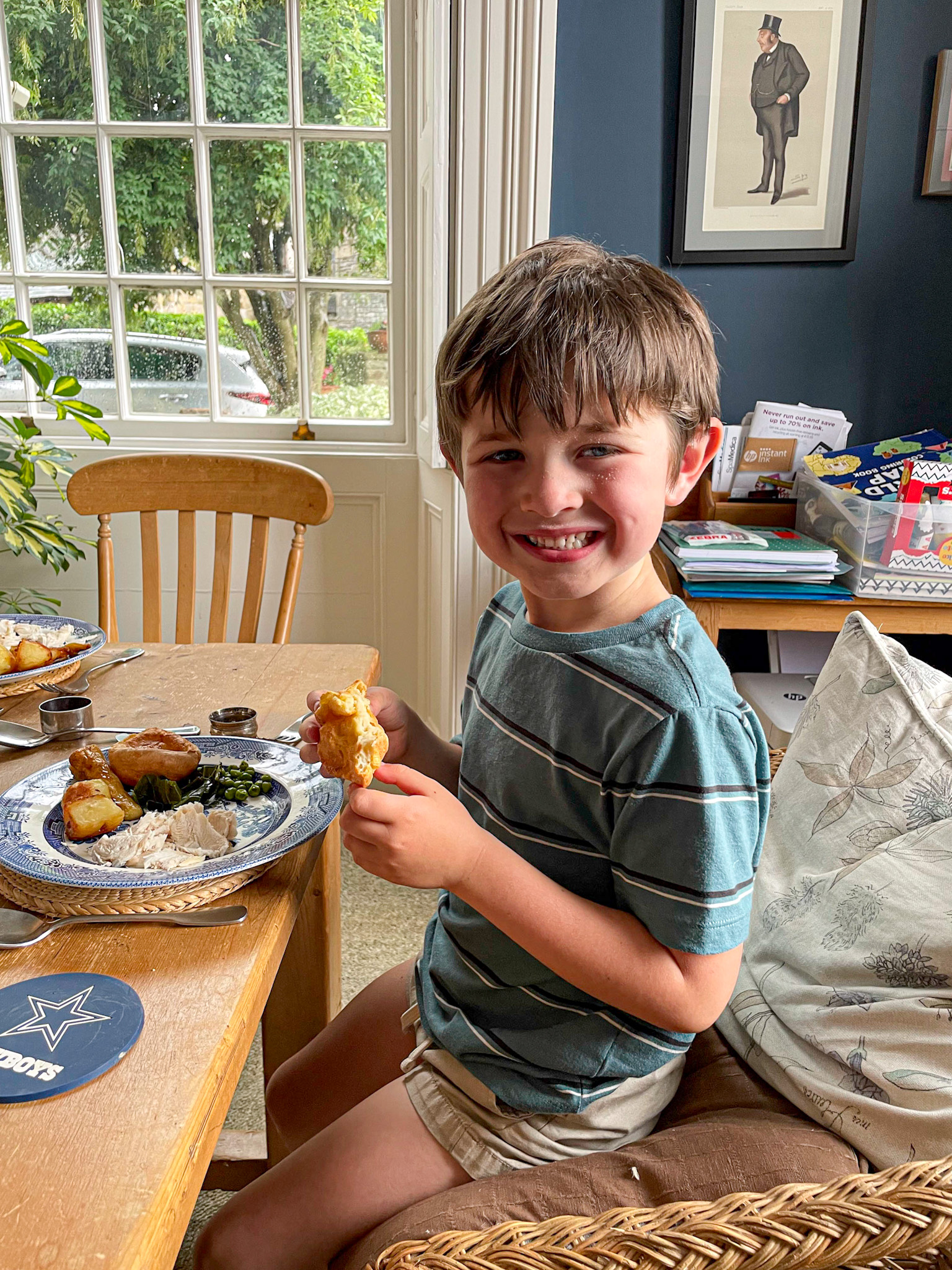 Smiling boy in a striped shirt eating a meal at a dining table in a cozy, light-filled room.