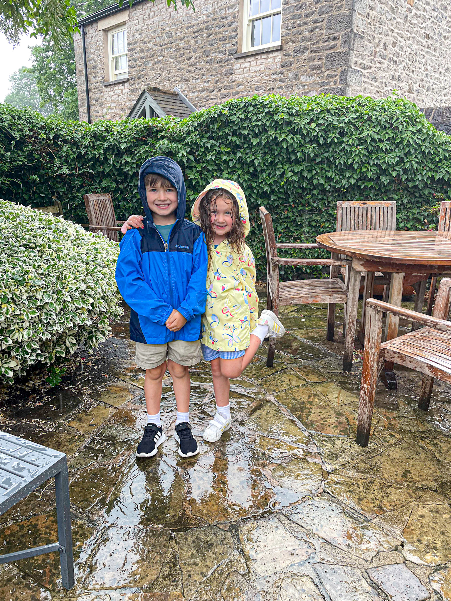 Two smiling kids in raincoats stand on a wet patio with outdoor furniture and greenery behind them.