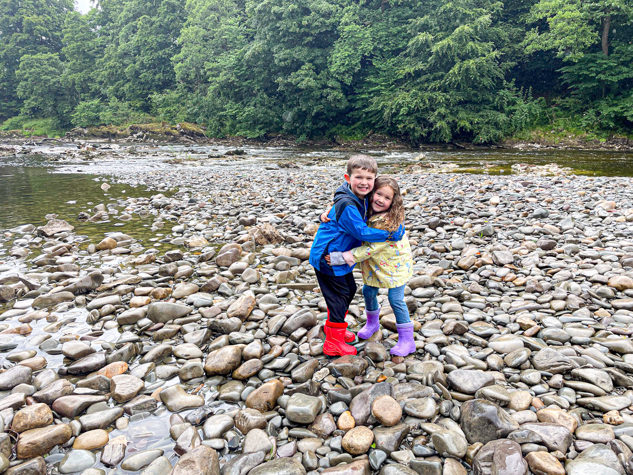 Two smiling children in colorful boots hug while standing on a rocky riverbank of the River Lune with trees in the background.