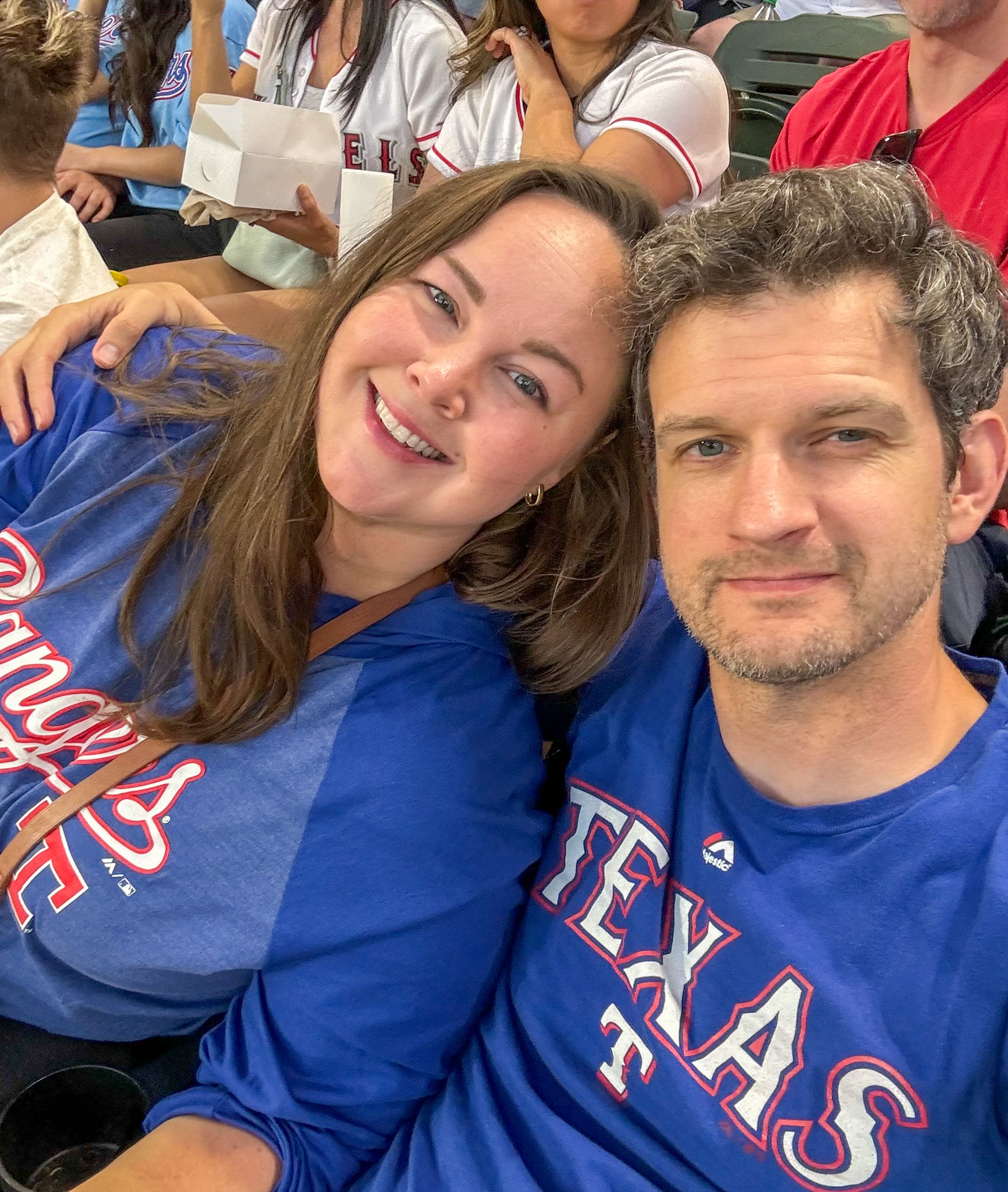 A smiling couple in Texas Rangers shirts pose for a selfie at a baseball game.